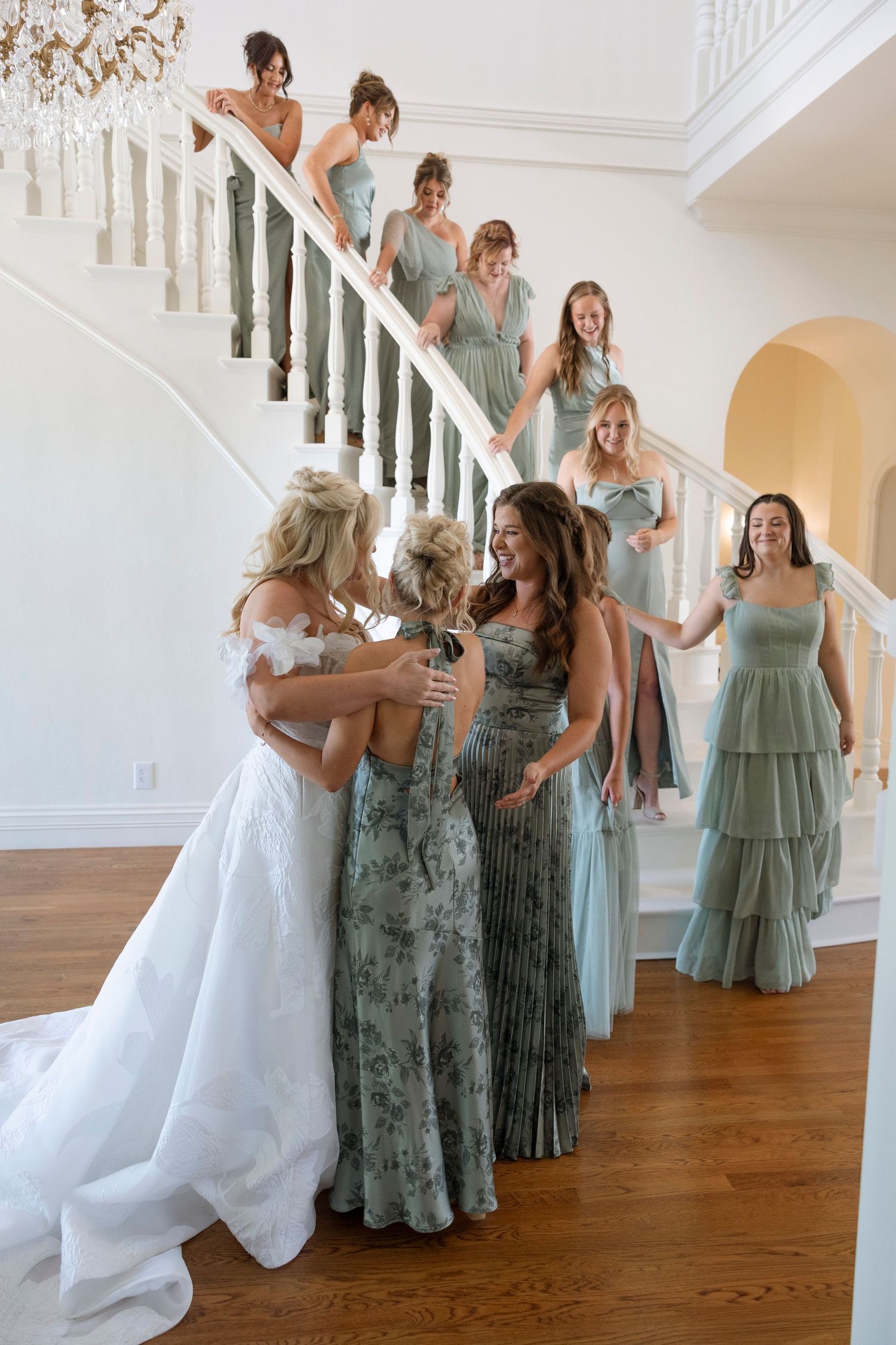 Wedding party in sage green dresses poses gracefully in a bright white room with hardwood floors.