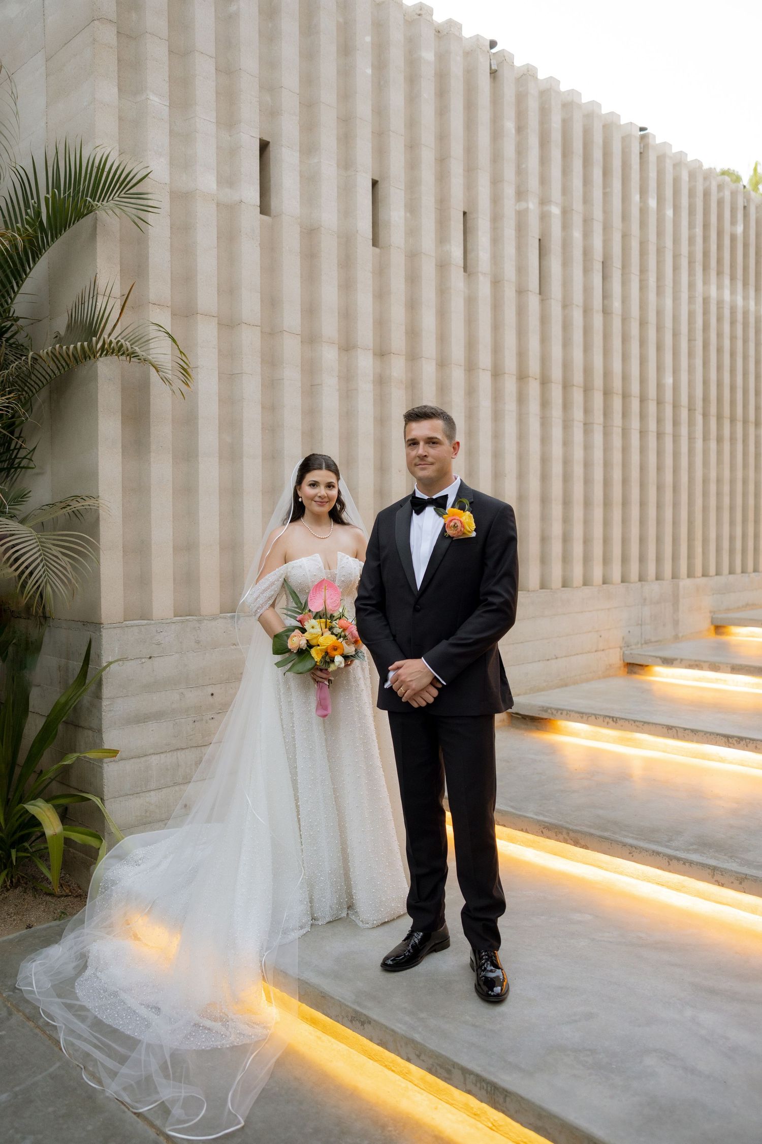 Bride and groom pose on illuminated steps beside textured stone wall, bride holds colorful bouquet.