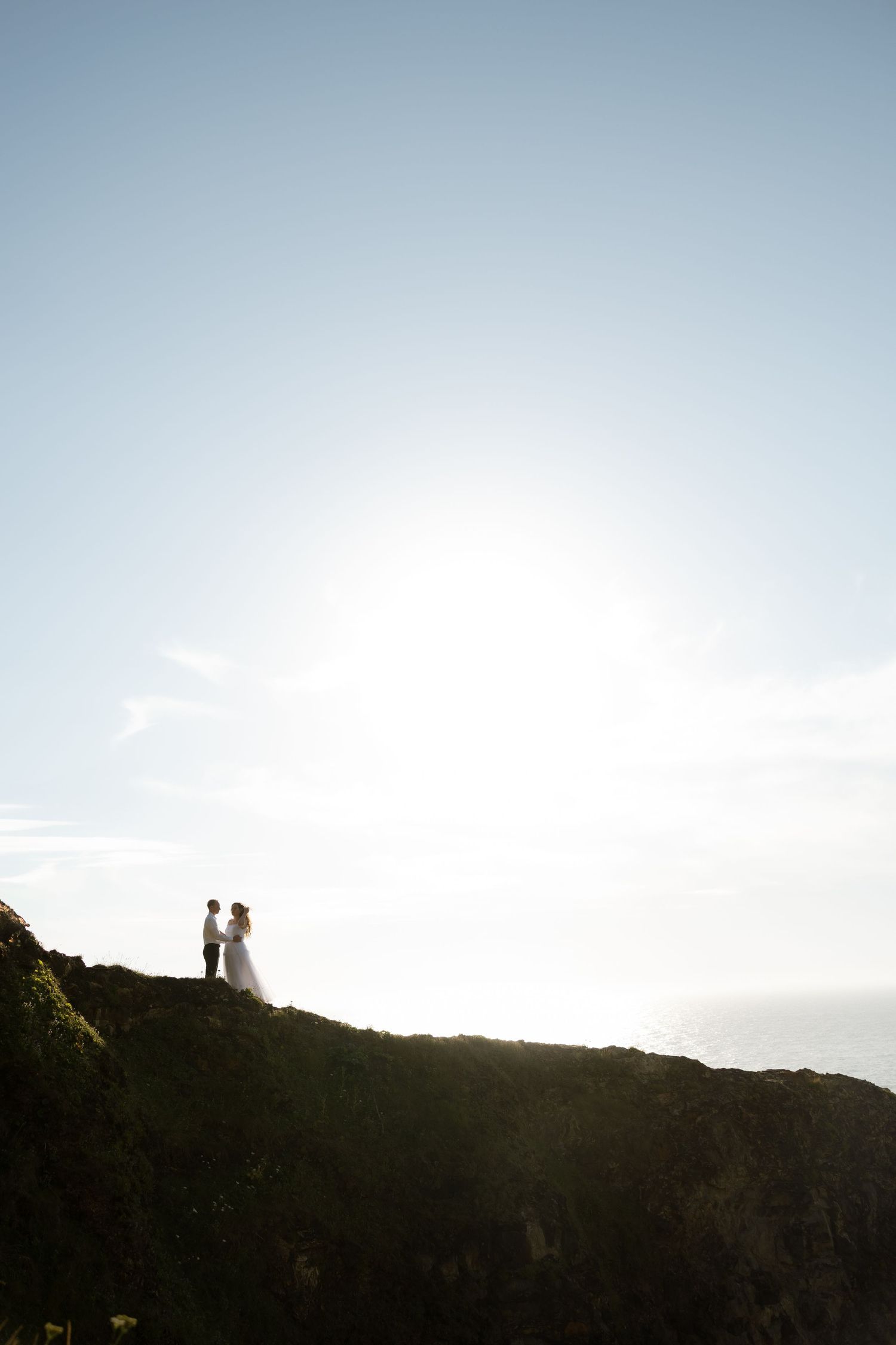 Silhouetted figures stand on a clifftop against a bright sky during a sunset wedding shoot.