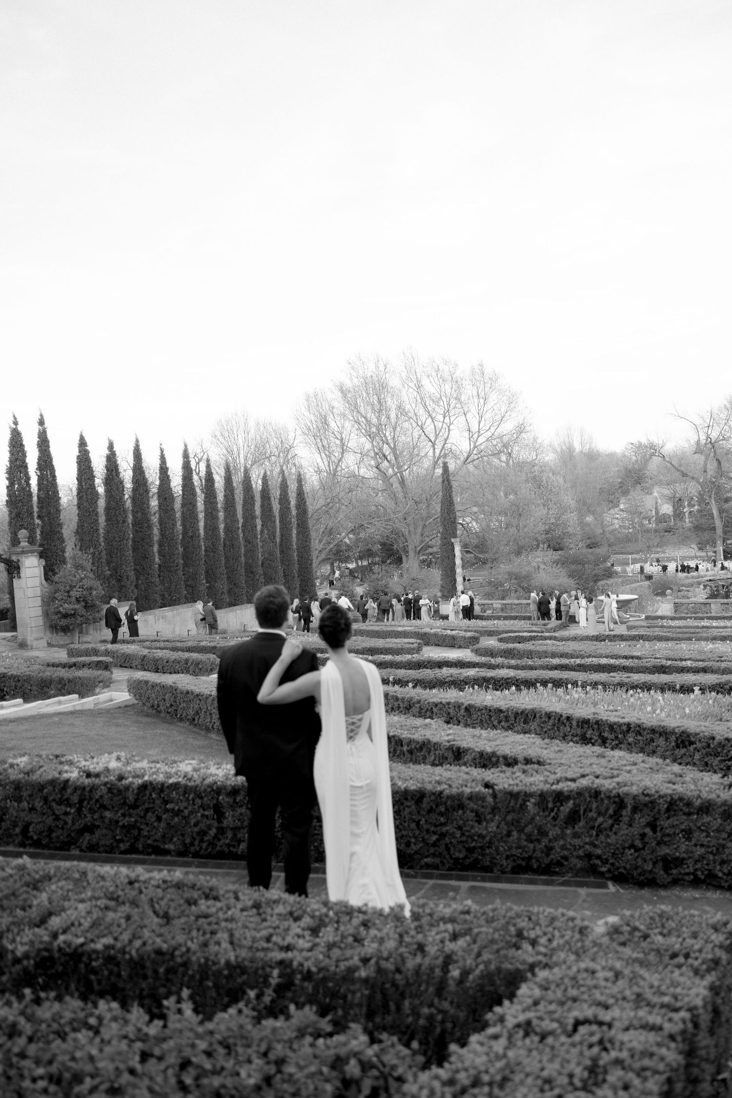Black and white photo of bride and groom overlooking formal garden with cypress trees.