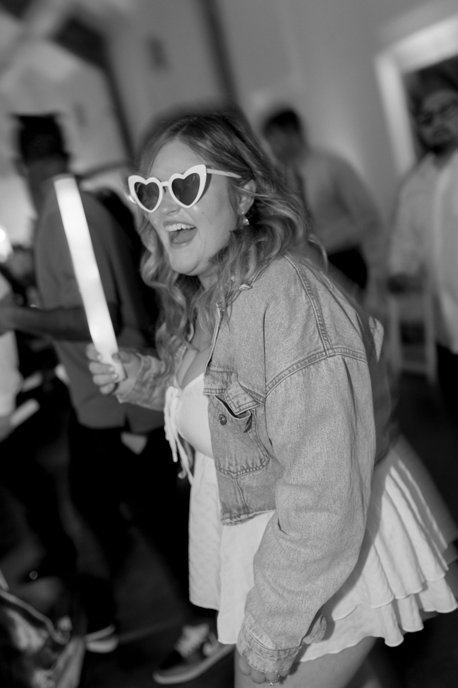 Dancing guest wearing heart-shaped sunglasses and denim jacket holds a light stick at wedding reception.