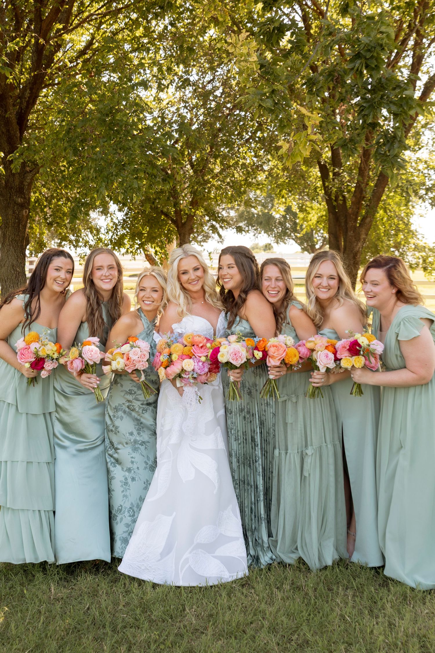 Wedding party wearing sage green bridesmaid dresses holding colorful bouquets pose together outdoors under trees.