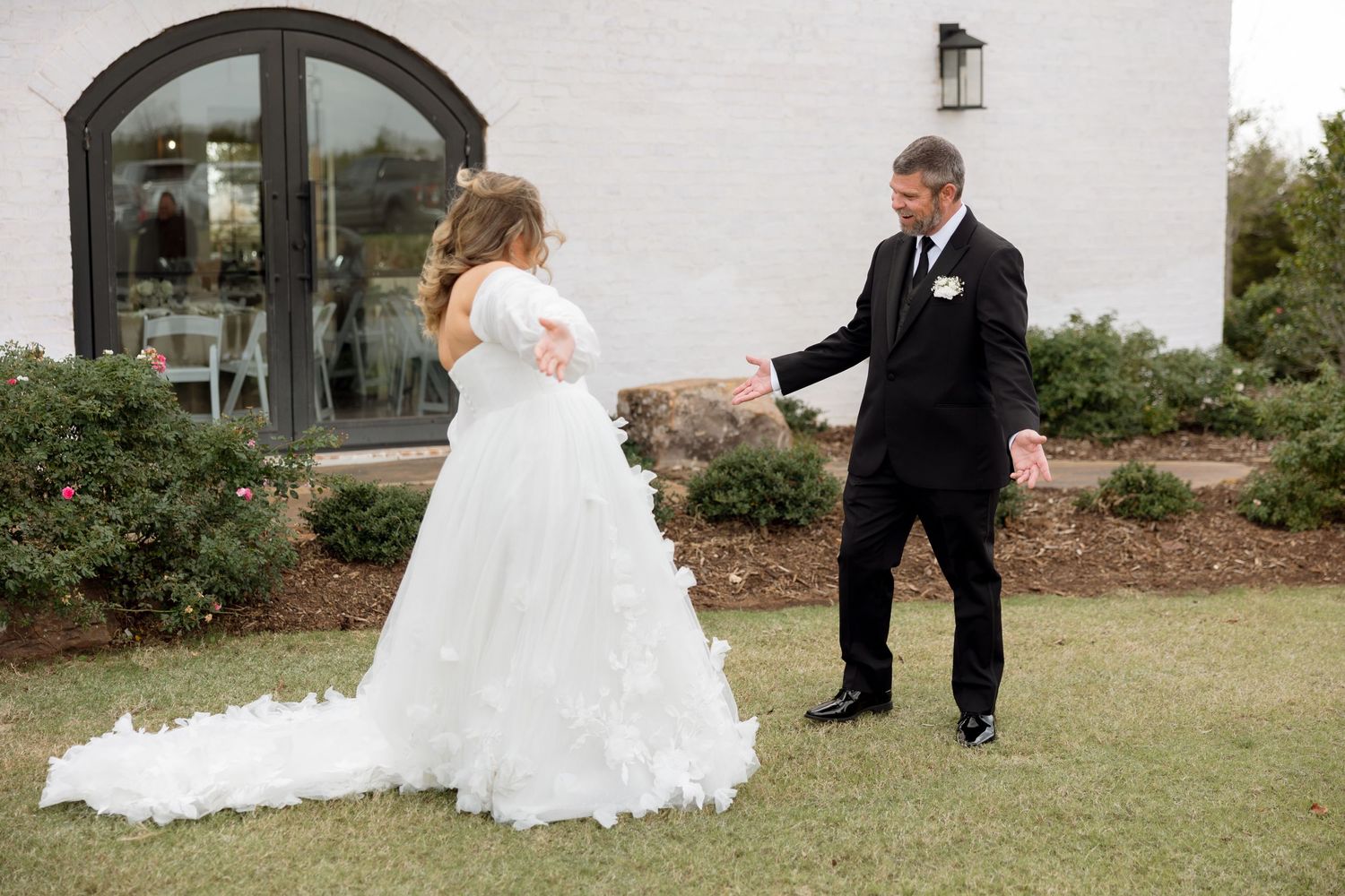Wedding couple shares candid first look moment on outdoor lawn near white building with arched windows.
