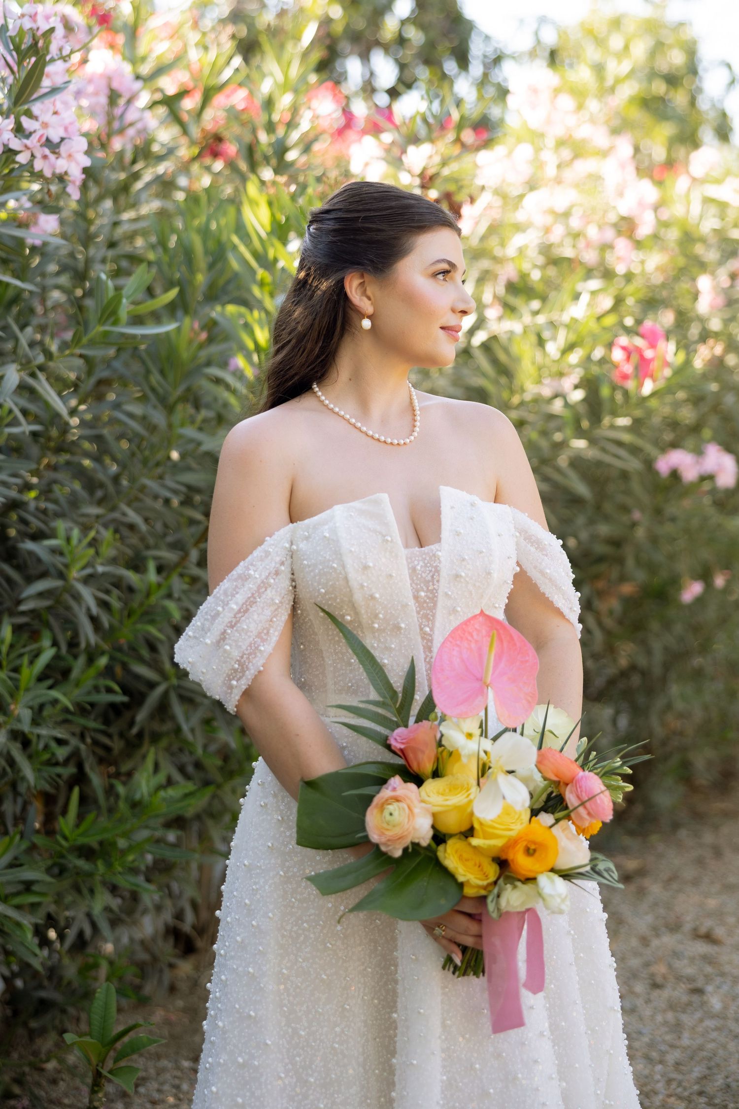 A bride in an off-shoulder pearl gown holds a tropical bouquet among blooming garden flowers.