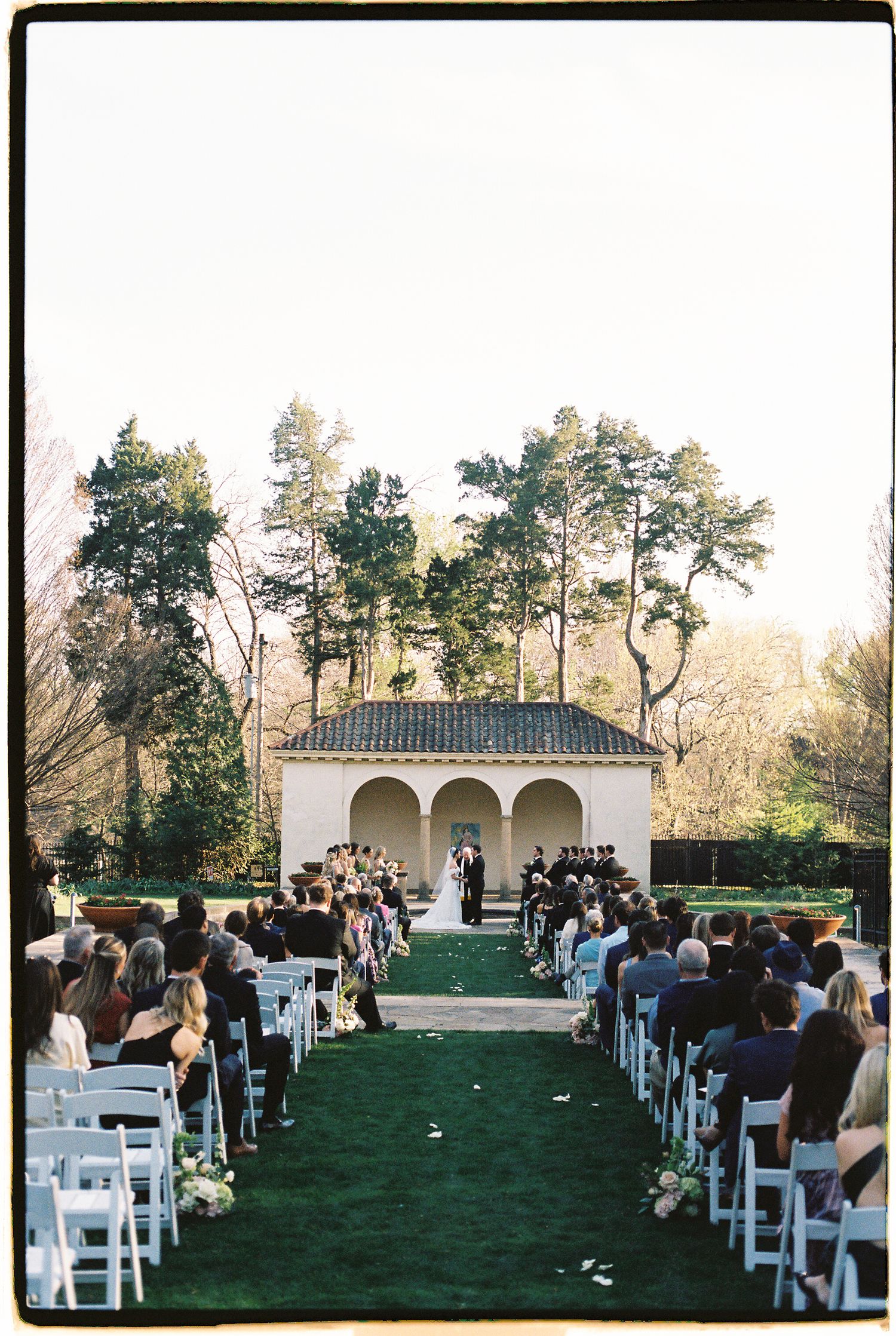 Elegant outdoor wedding ceremony on green lawn with white chairs and a charming Spanish-style pavilion backdrop.
