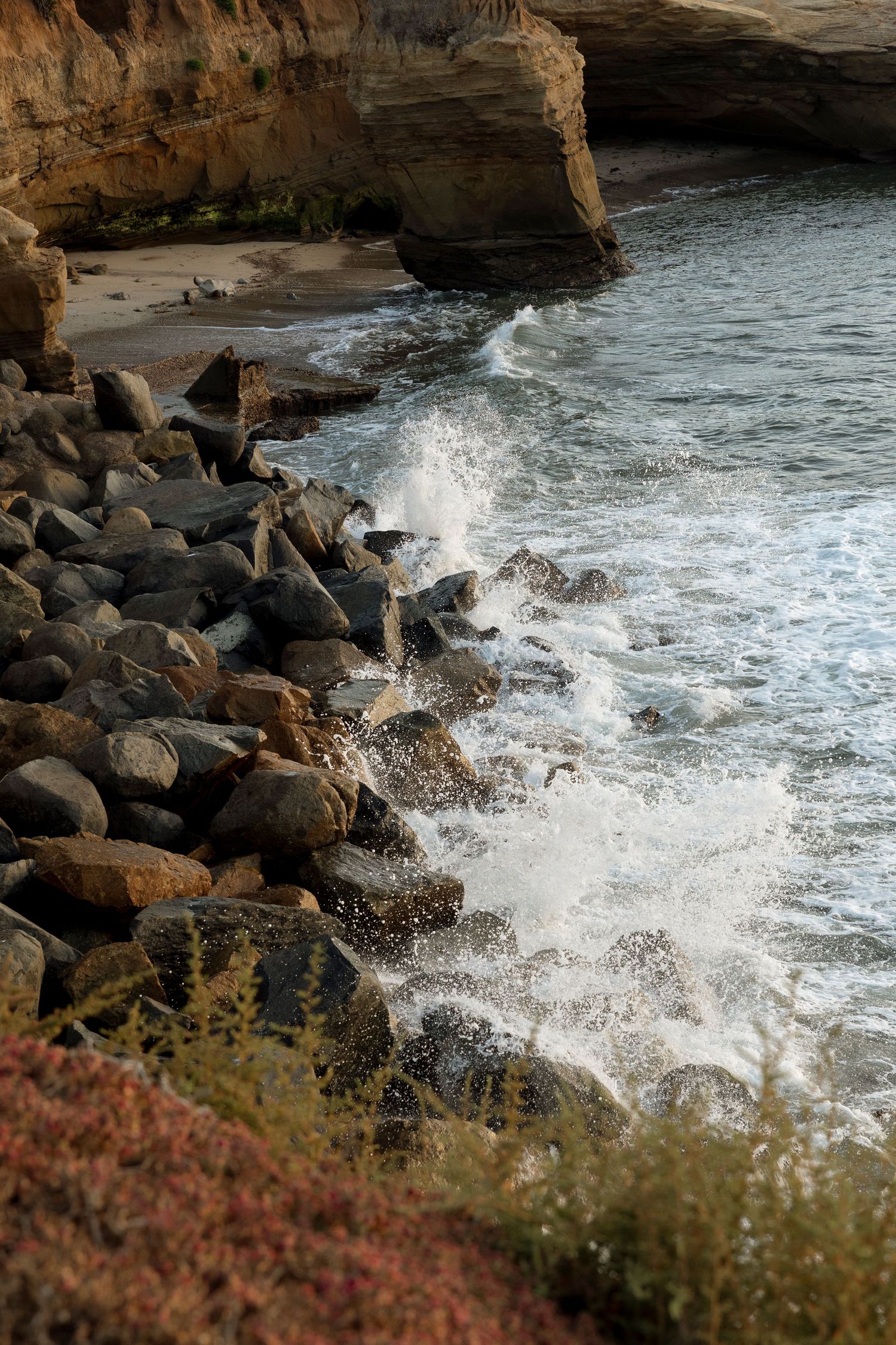 Ocean waves crash dramatically against rocky coastal cliffs during sunset.