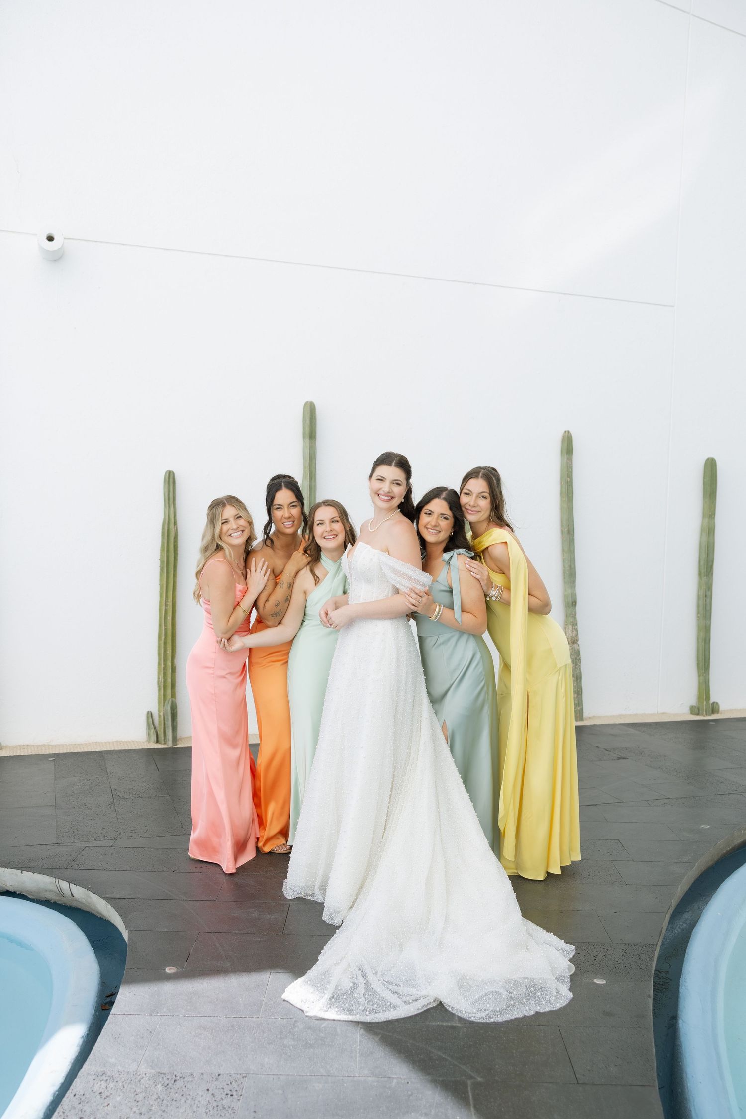 Bride in white gown poses with five bridesmaids in colorful mismatched dresses near a pool with cacti backdrop.