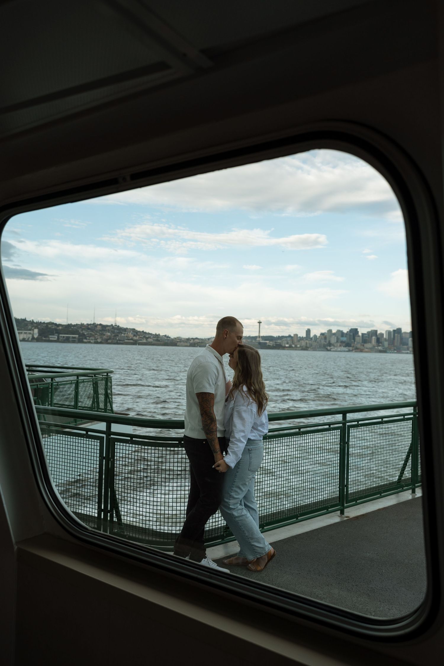 Ferry window frames couple on deck with Seattle skyline in background.