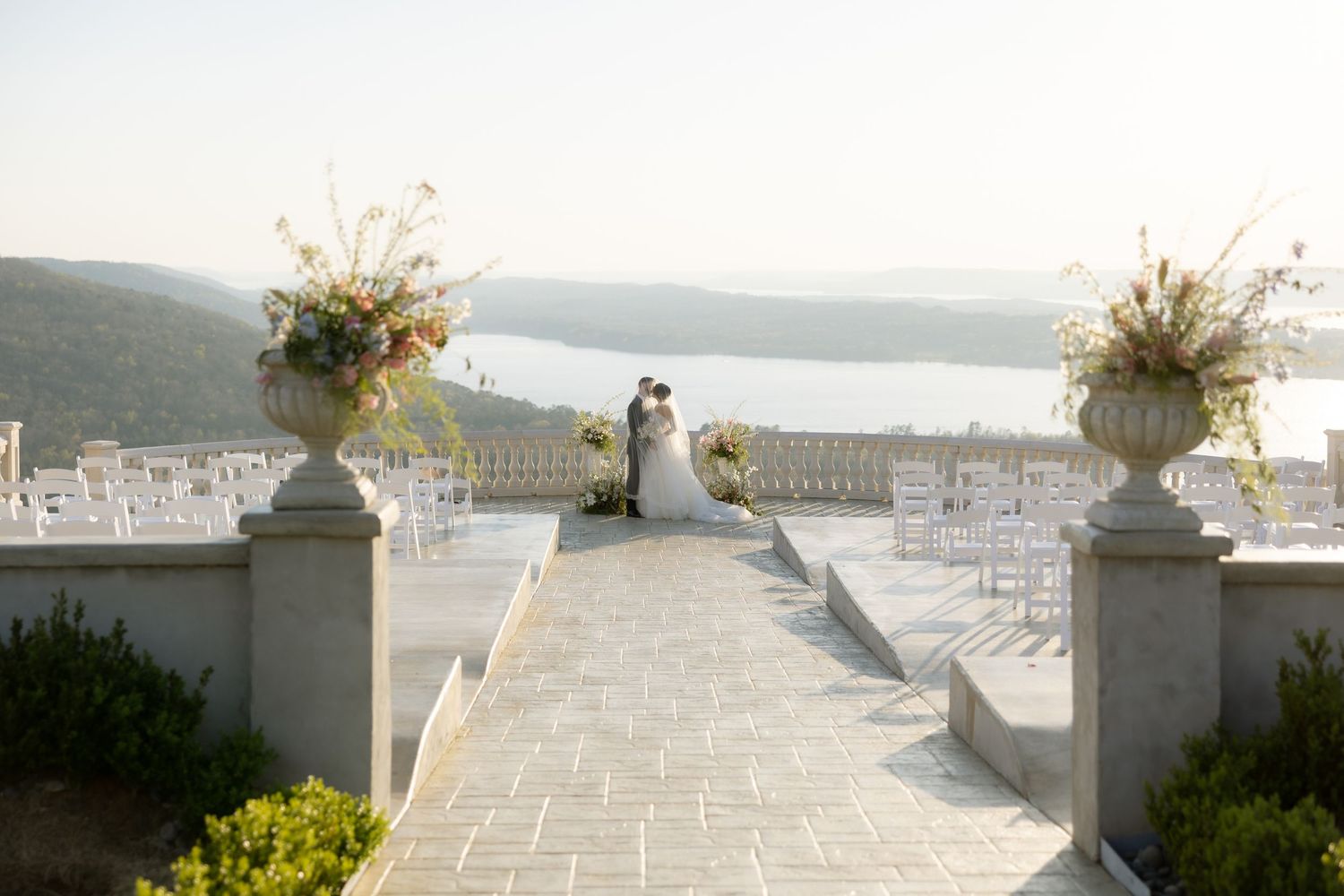 Elegant outdoor wedding ceremony on a stone terrace with floral urns, overlooking a scenic river valley.