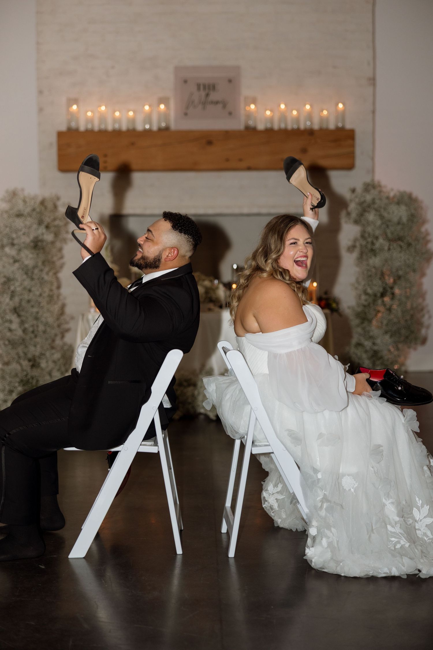 Wedding couple playing the shoe game while sitting back-to-back on white chairs at their reception.