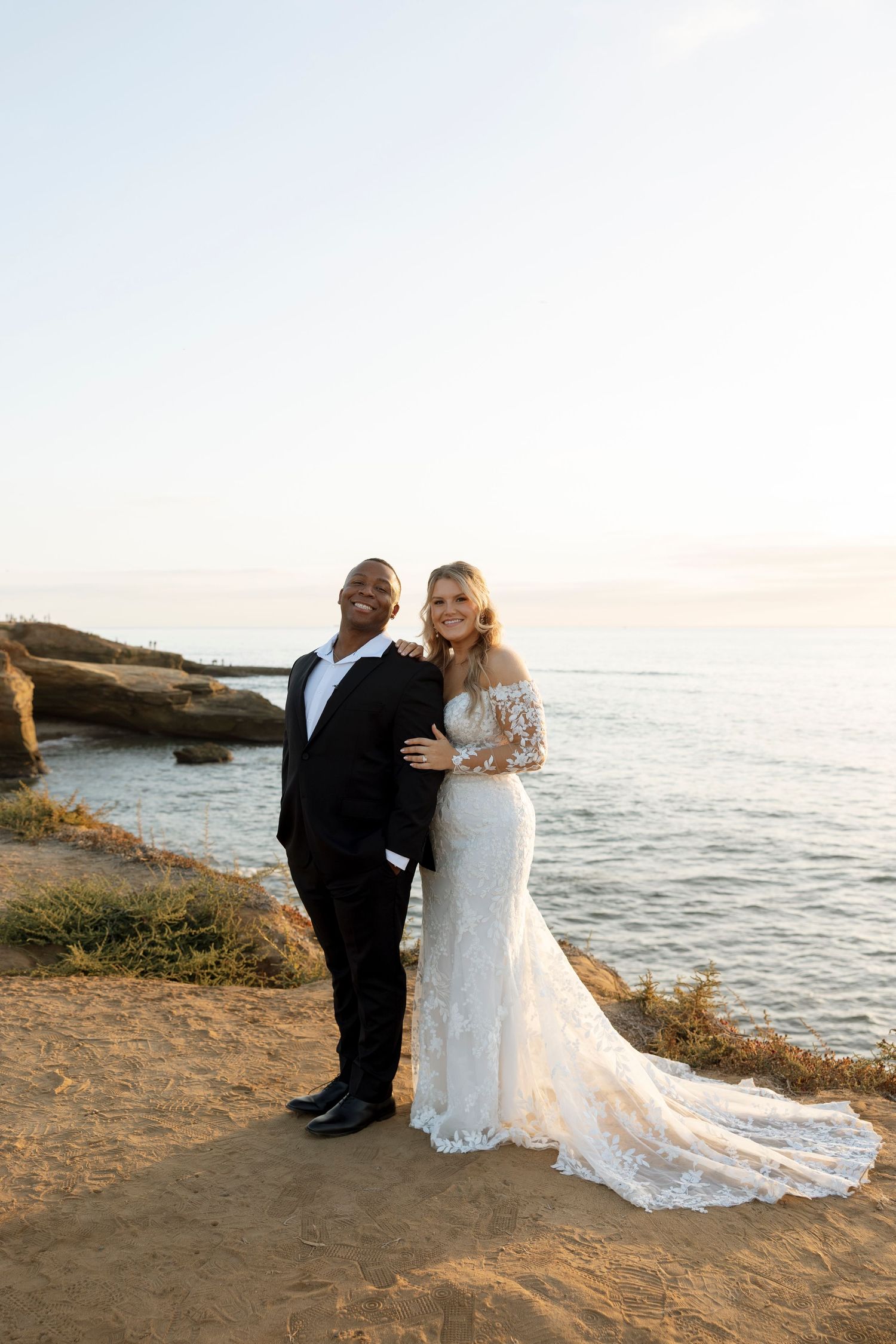 Couple in formal attire stands together on coastal cliffs overlooking the ocean at sunset.