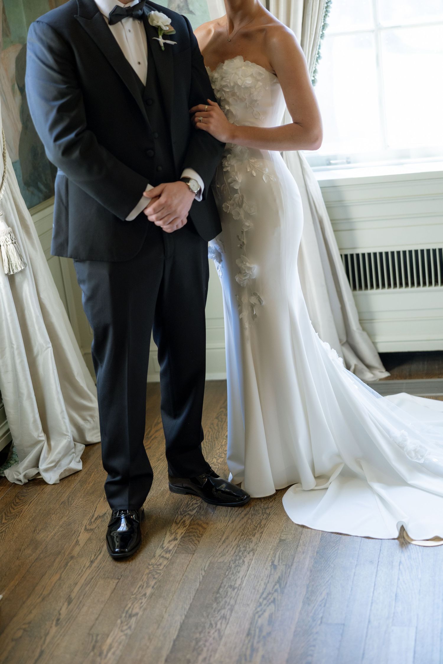 Close-up of bride in strapless lace gown and groom in dark tuxedo standing by window indoors.