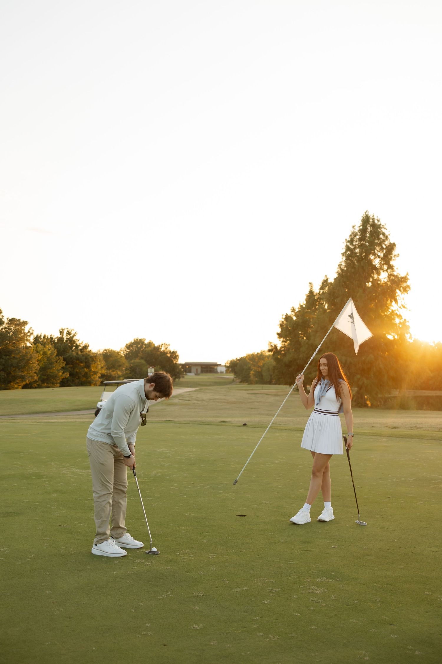 Two people golfing together on a putting green with a sunset-lit pine tree in background.