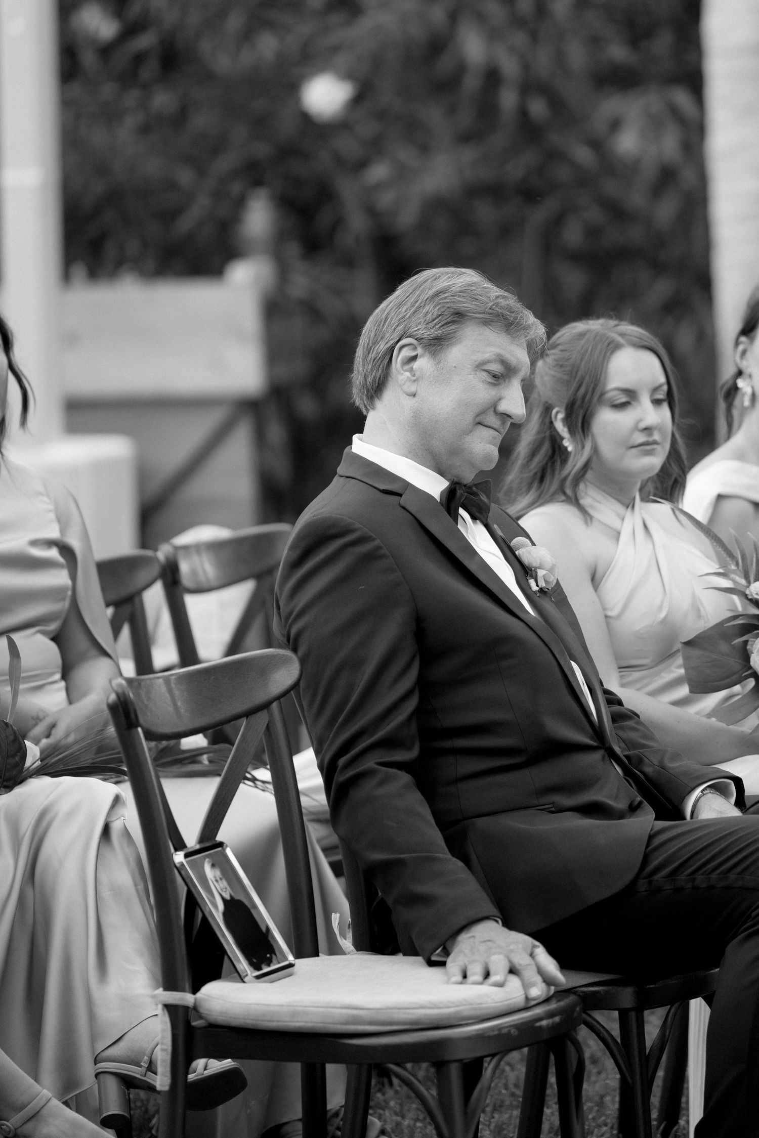 A man in a tuxedo sits smiling at an outdoor wedding ceremony in black and white photography.
