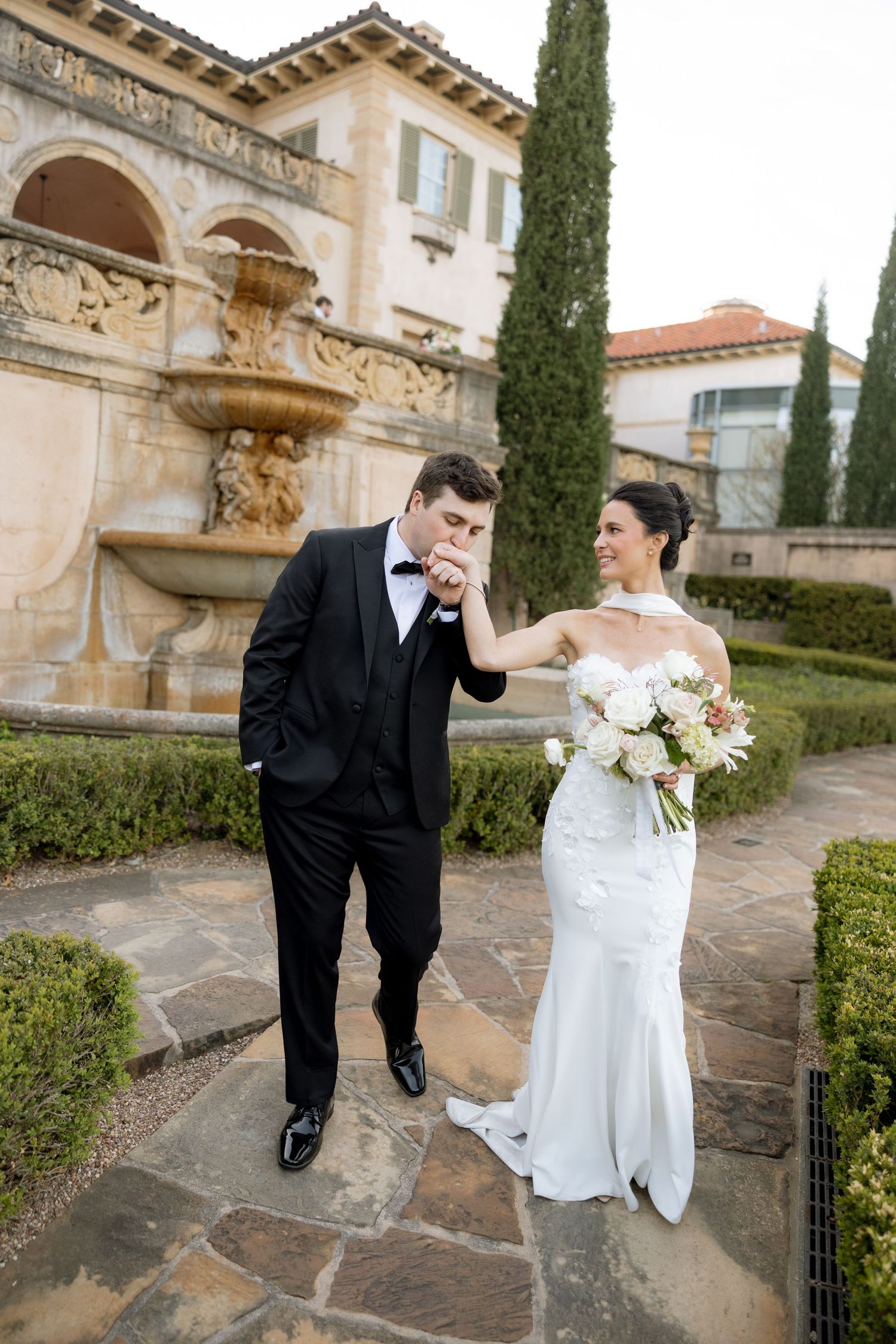 Groom in tuxedo kisses bride's hand as she smiles holding bouquet near ornate fountain at estate wedding.