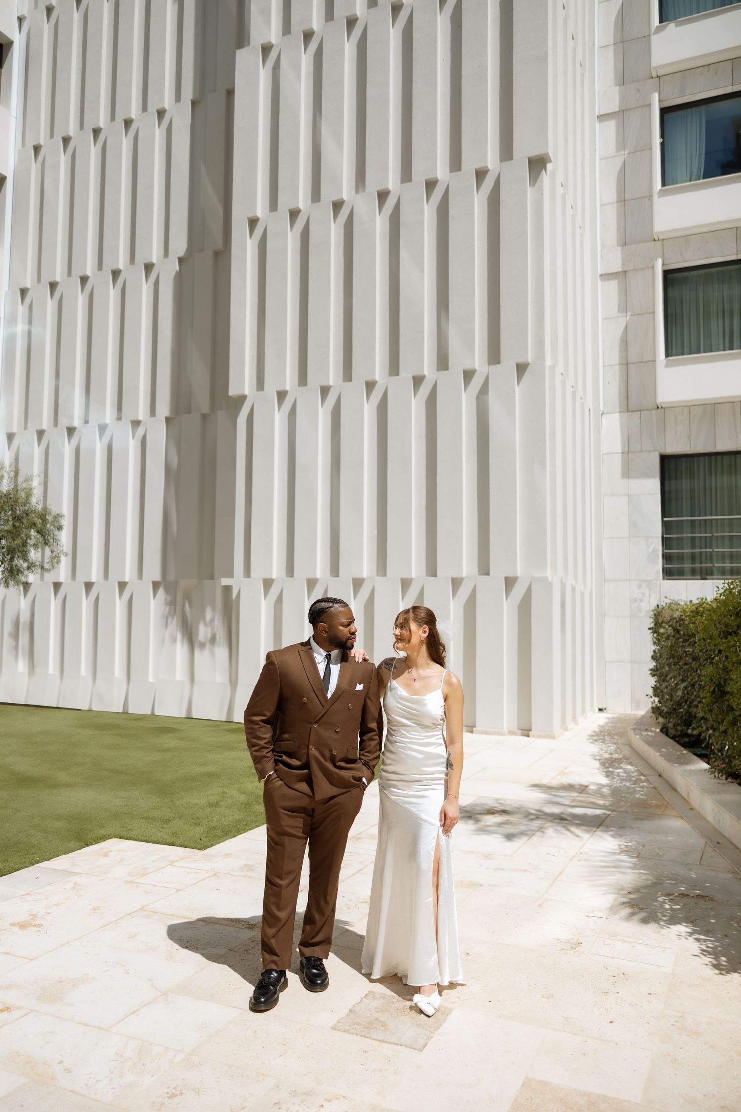 A bride and groom stand together by a geometric white wall during their wedding photoshoot.