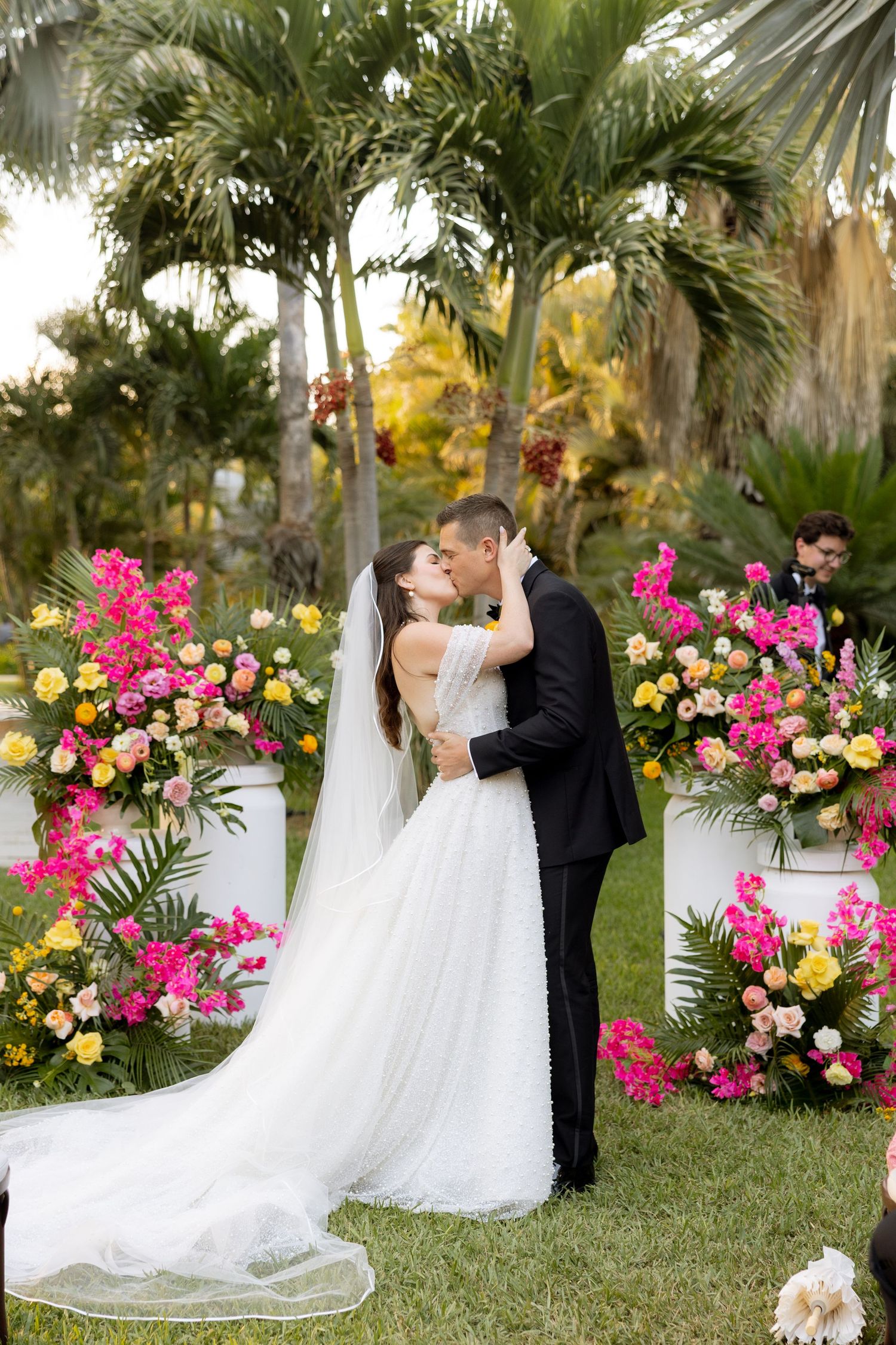 A bride and groom share their first kiss at an outdoor wedding ceremony surrounded by vibrant pink and yellow floral arrangements.