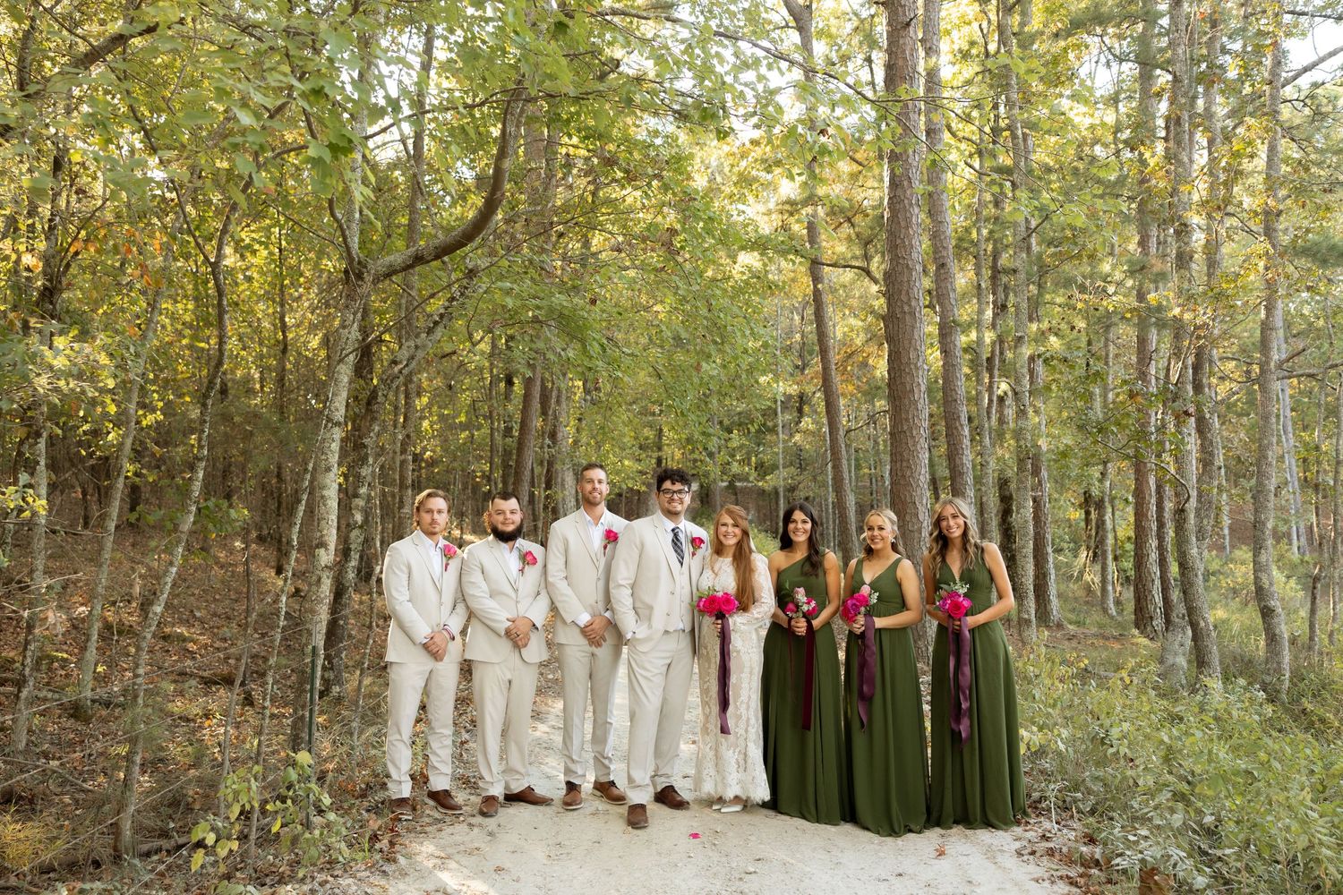 Wedding party in white and olive green attire poses together on a forest path during golden hour.