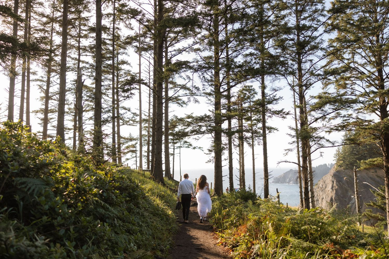 Couple walks hand in hand on forest trail overlooking coastal cliffs.