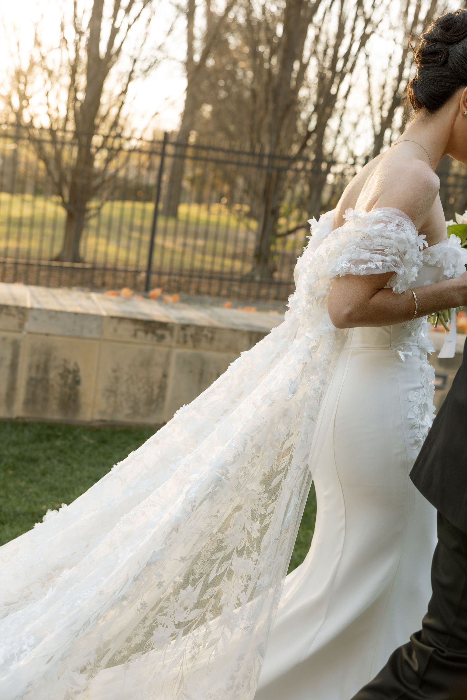 Bride in off-shoulder floral wedding gown with flowing lace train walking outdoors at golden hour.
