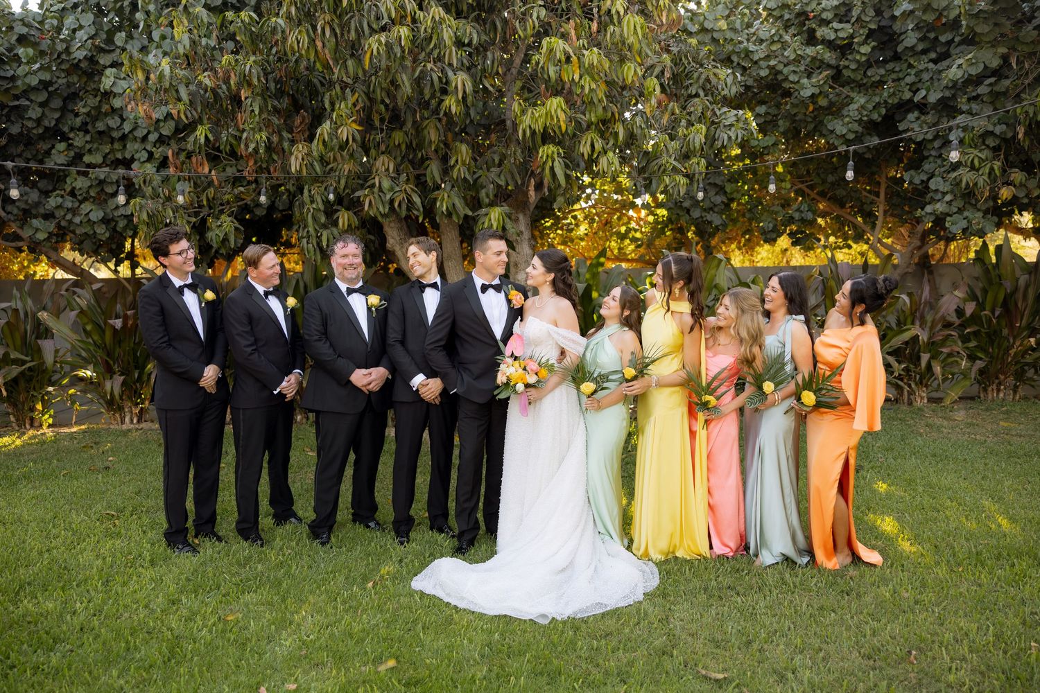 Wedding party poses outdoors with groomsmen in black tuxedos and bridesmaids in colorful yellow and green gowns.