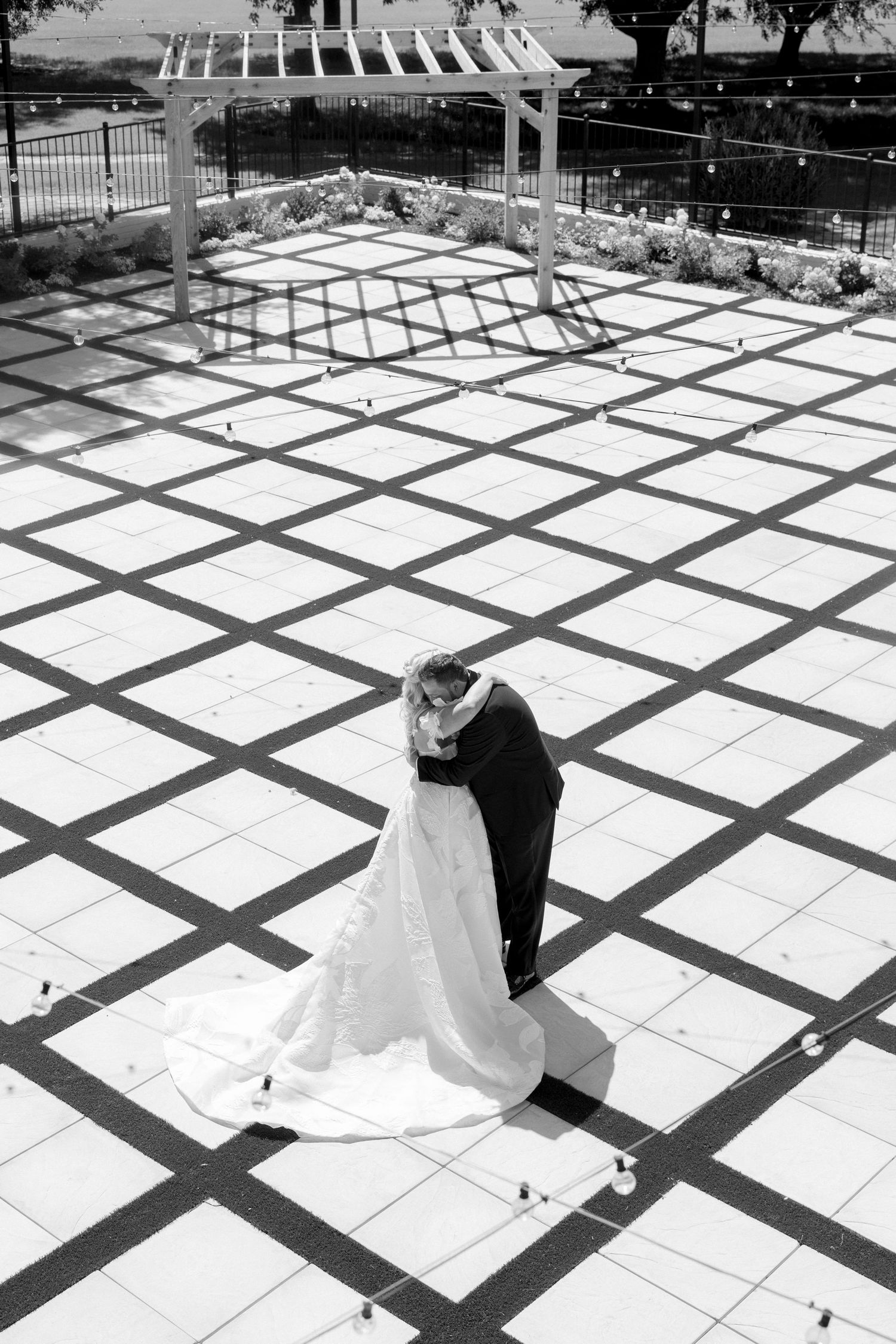 A black and white photograph of a couple dancing on a geometric patterned patio under a pergola.