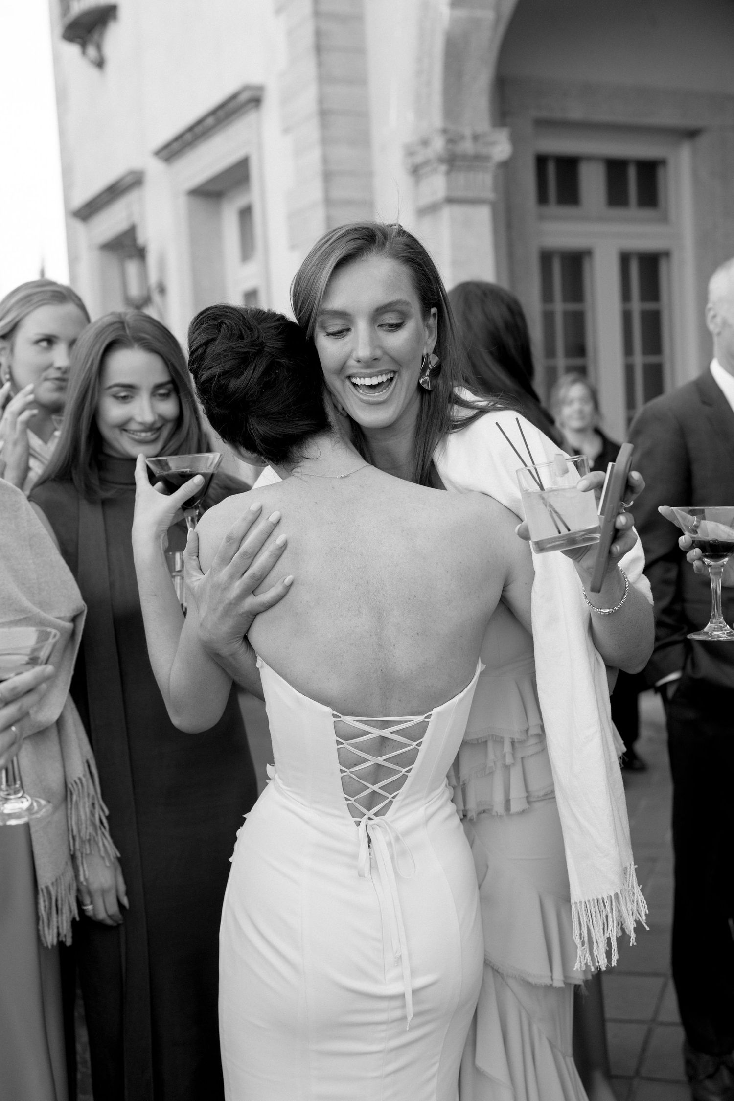 Black and white photo of bride in backless lace-up gown embracing guest at outdoor cocktail hour.