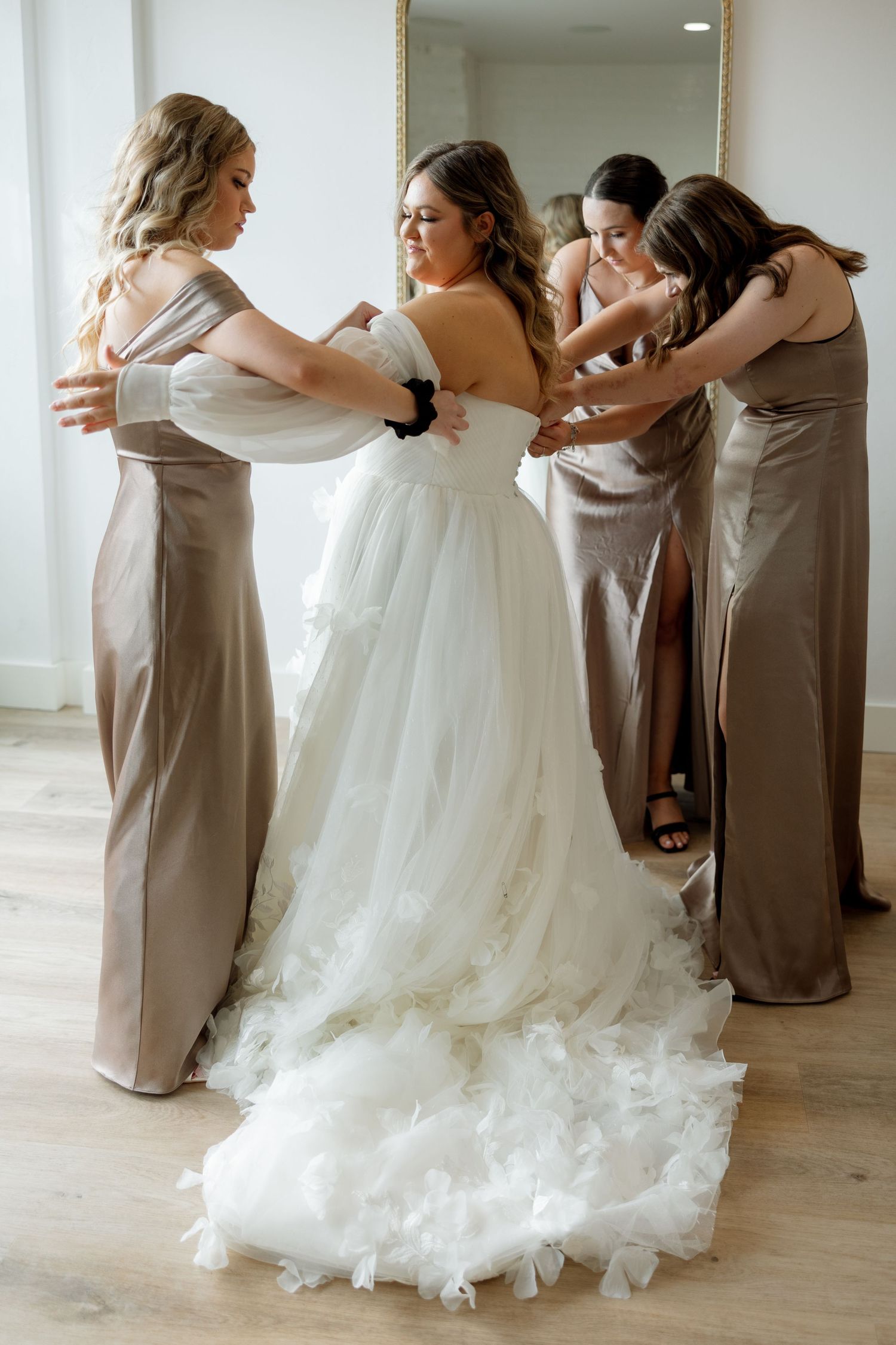 Bridesmaids in taupe dresses help the bride get ready in her flowing white gown.