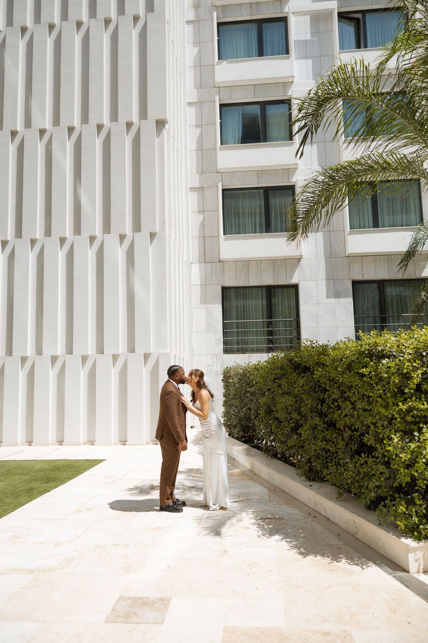 A couple shares an intimate moment against a modern white architectural facade with palm trees.
