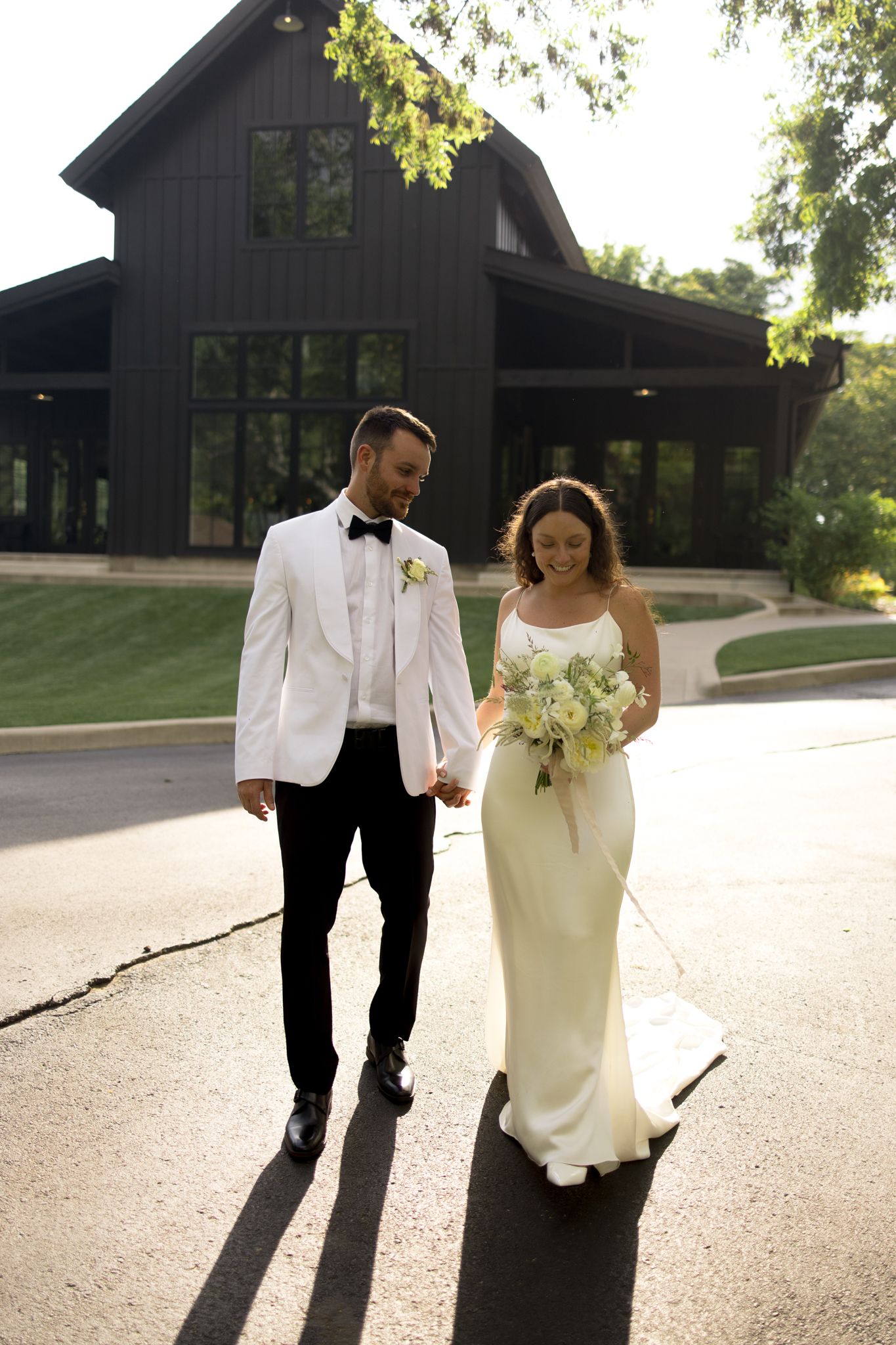 Elegant couple in wedding attire walking together outside a modern black barn venue.