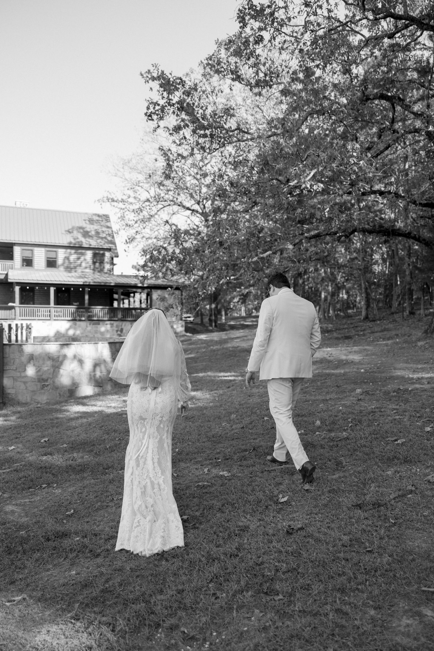 Black and white photo of a couple walking away from camera across a lawn near a building.
