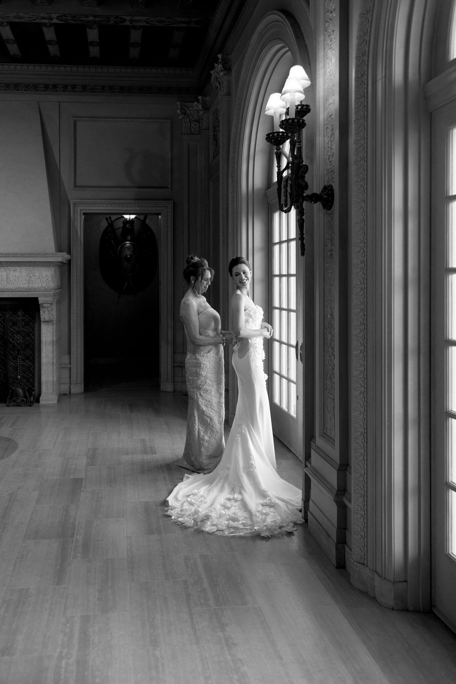 Two brides in white wedding gowns stand by arched windows in a grand historic mansion in black and white photo.
