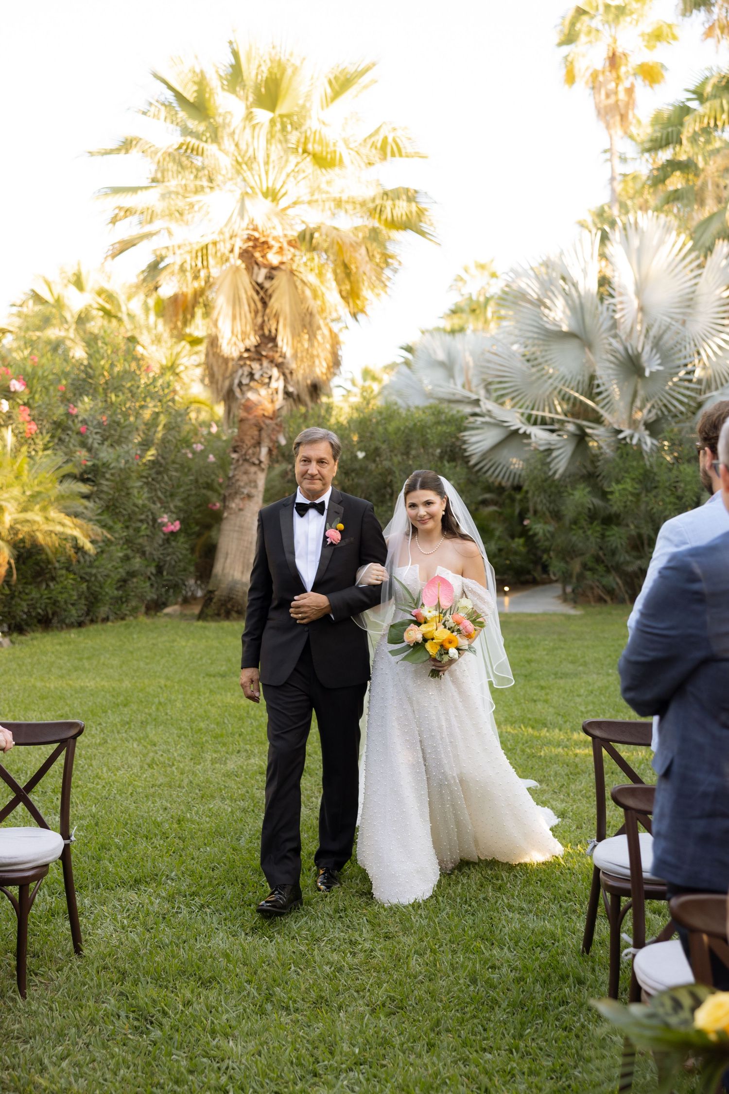 Bride in white gown walks down outdoor aisle with escort amid palm trees at sunset wedding ceremony.
