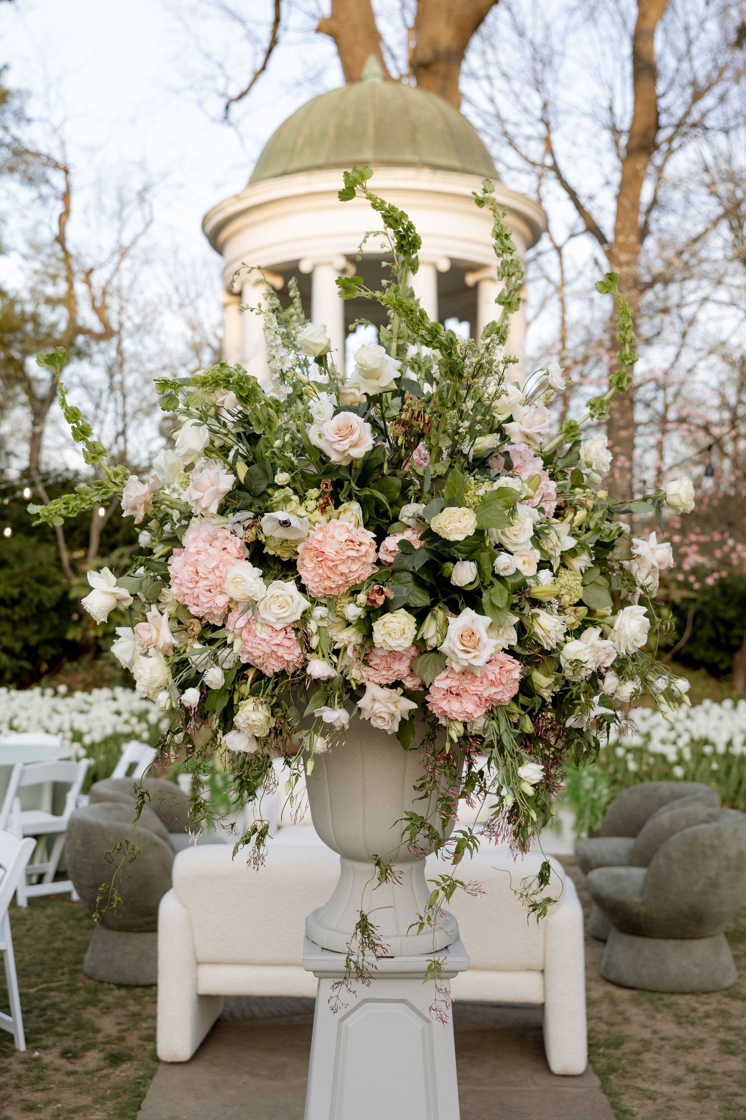 Large floral arrangement of pink hydrangeas and white roses in a white urn with a classical gazebo in the background.