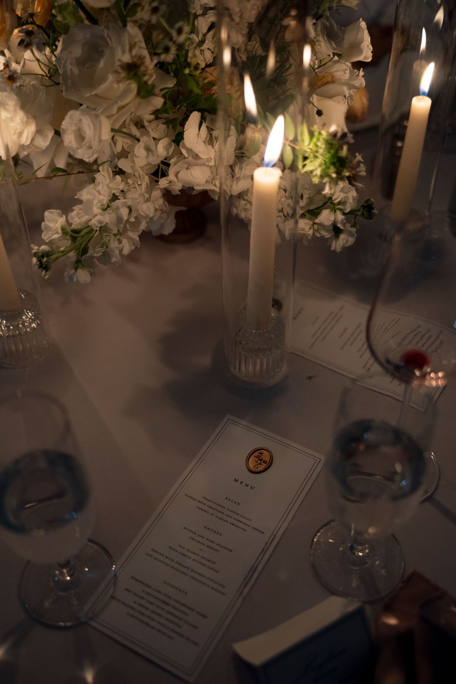 Elegant wedding reception table setting with white floral centerpieces, glowing candles and menu cards.