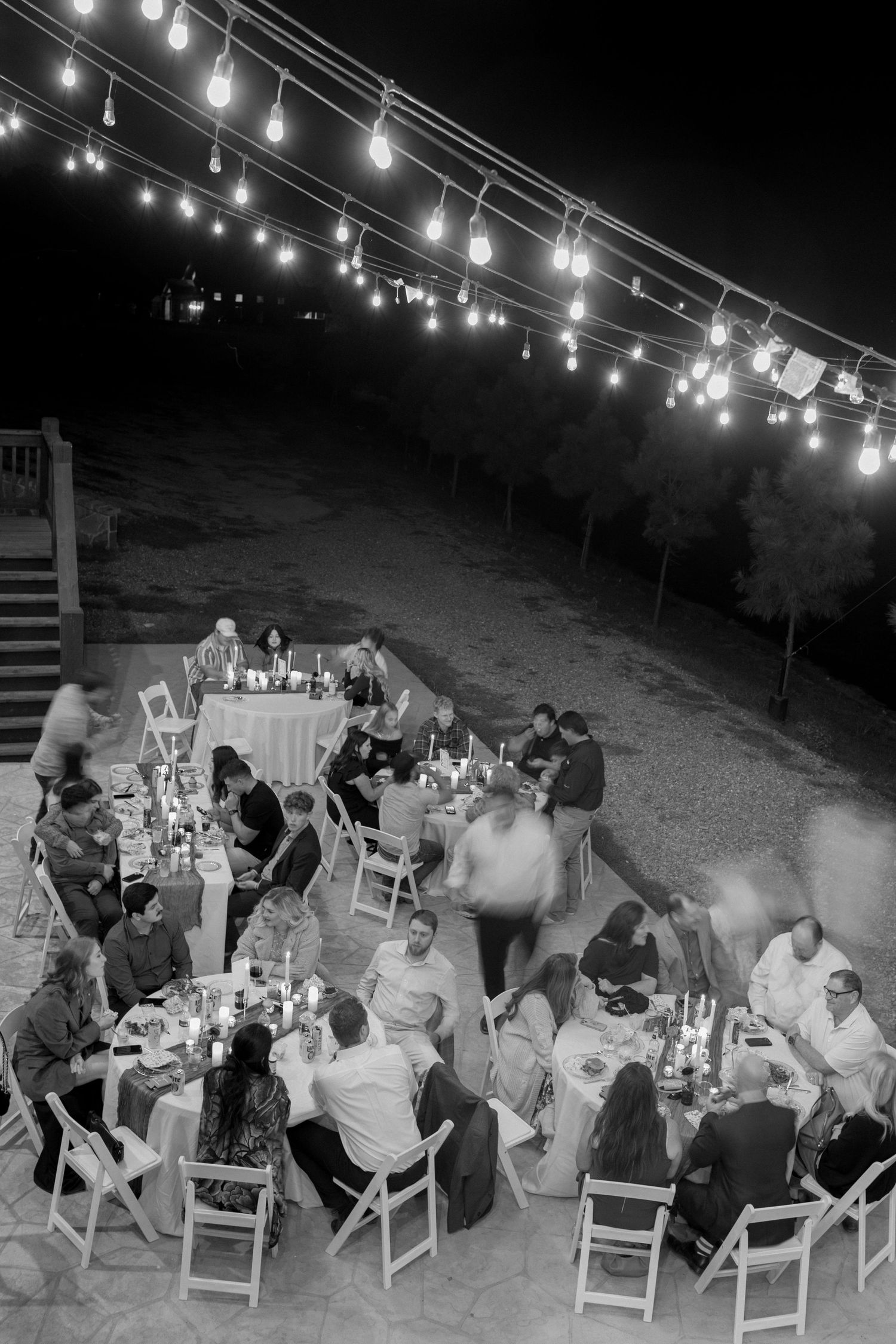 Black and white image of outdoor wedding reception with string lights and guests seated at tables.