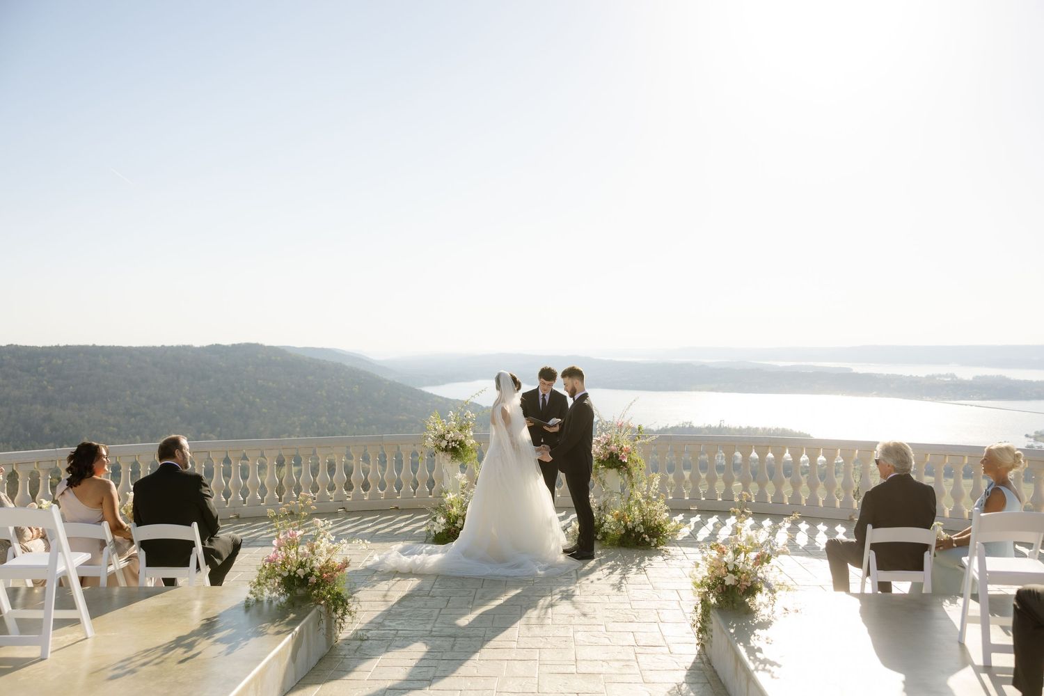 Outdoor wedding ceremony on a scenic terrace with mountain and river views, bride and groom at altar with guests seated.