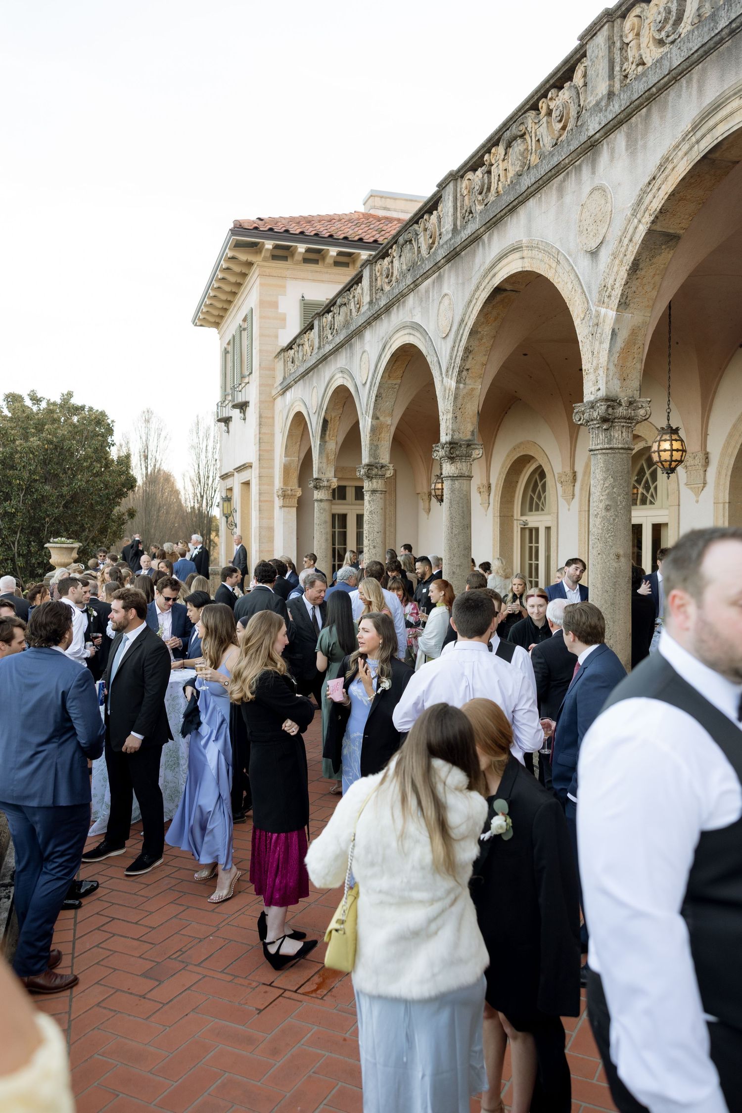 Elegantly dressed wedding guests mingle outside a grand Italian-style venue with arched colonnades.