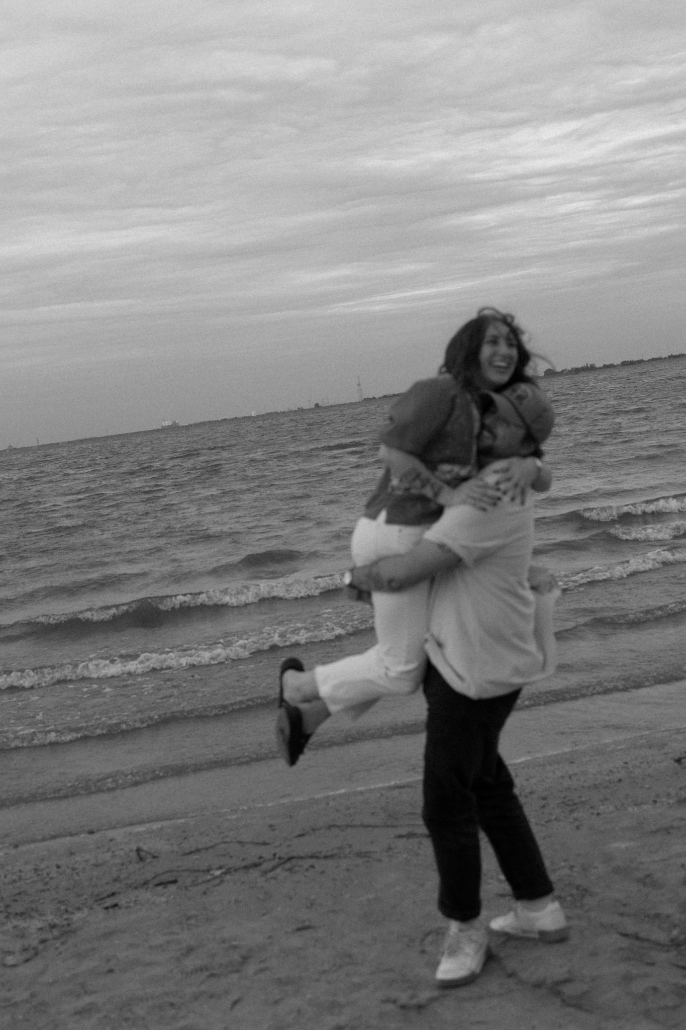 A playful couple embracing and laughing together on a beach at sunset in black and white.