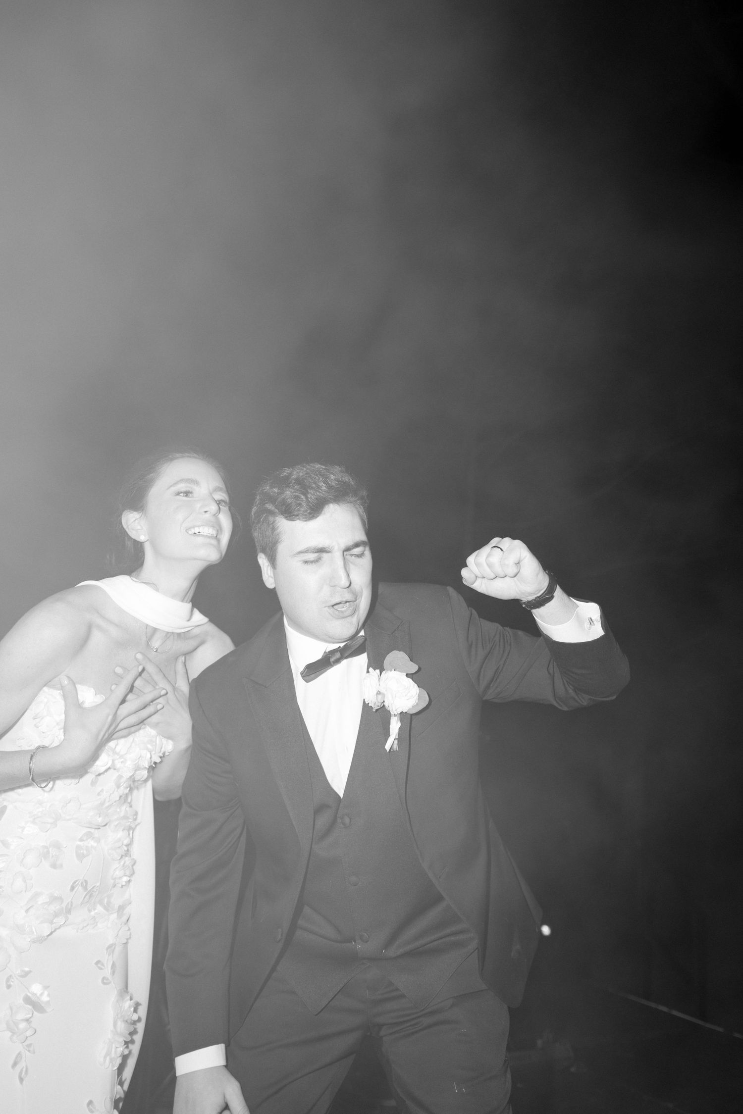 Black and white photo of bride and groom dancing joyfully on smoky dance floor at wedding reception.