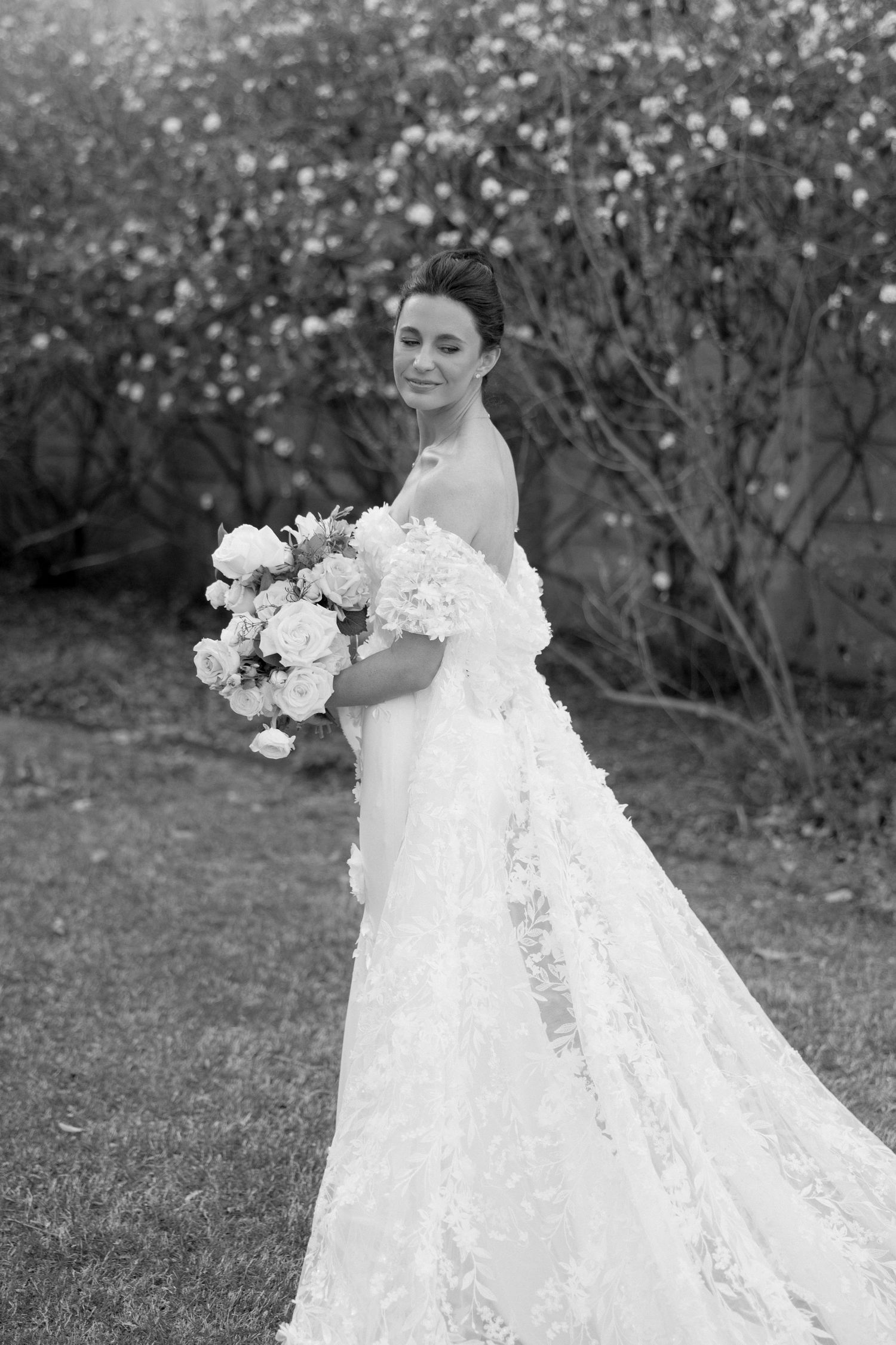Bride in floral lace gown holding bouquet of white peonies in garden setting, black and white.