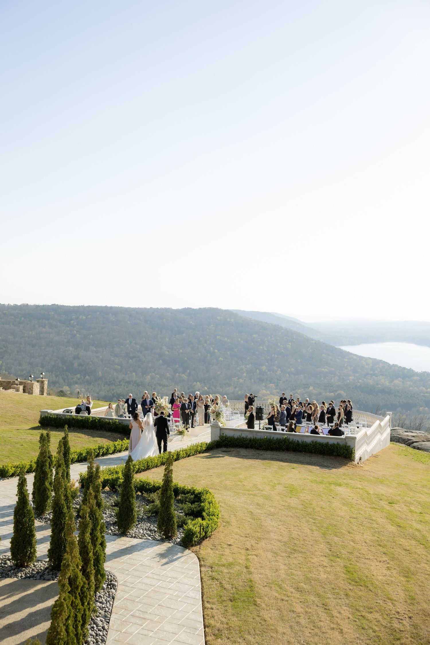 Outdoor wedding ceremony on a hilltop terrace overlooking a scenic river valley and forested mountains.