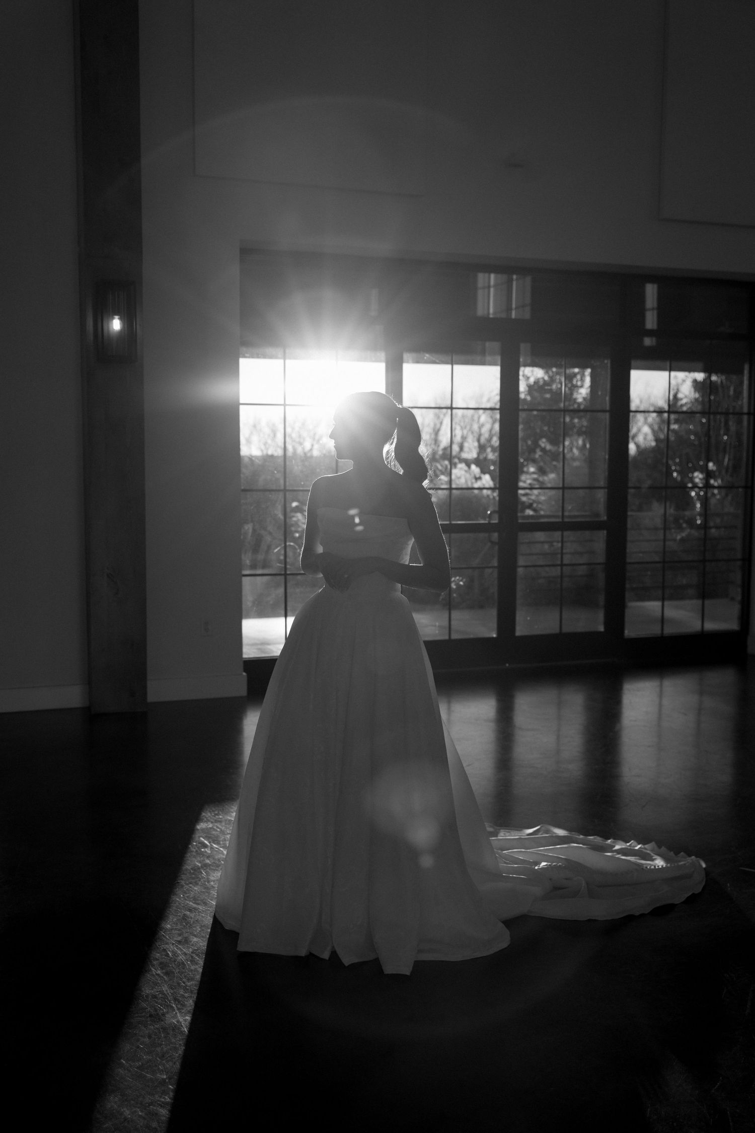 Black and white silhouette of a bride in a flowing wedding dress standing by window with dramatic backlighting.