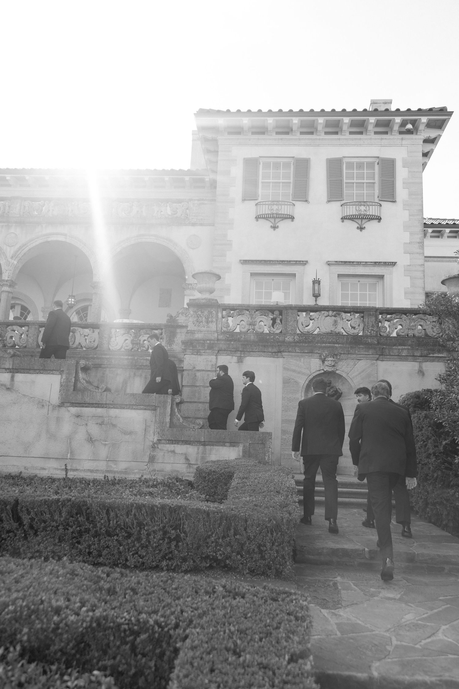 Black and white photo of groomsmen in suits ascending stone steps of grand estate wedding venue.
