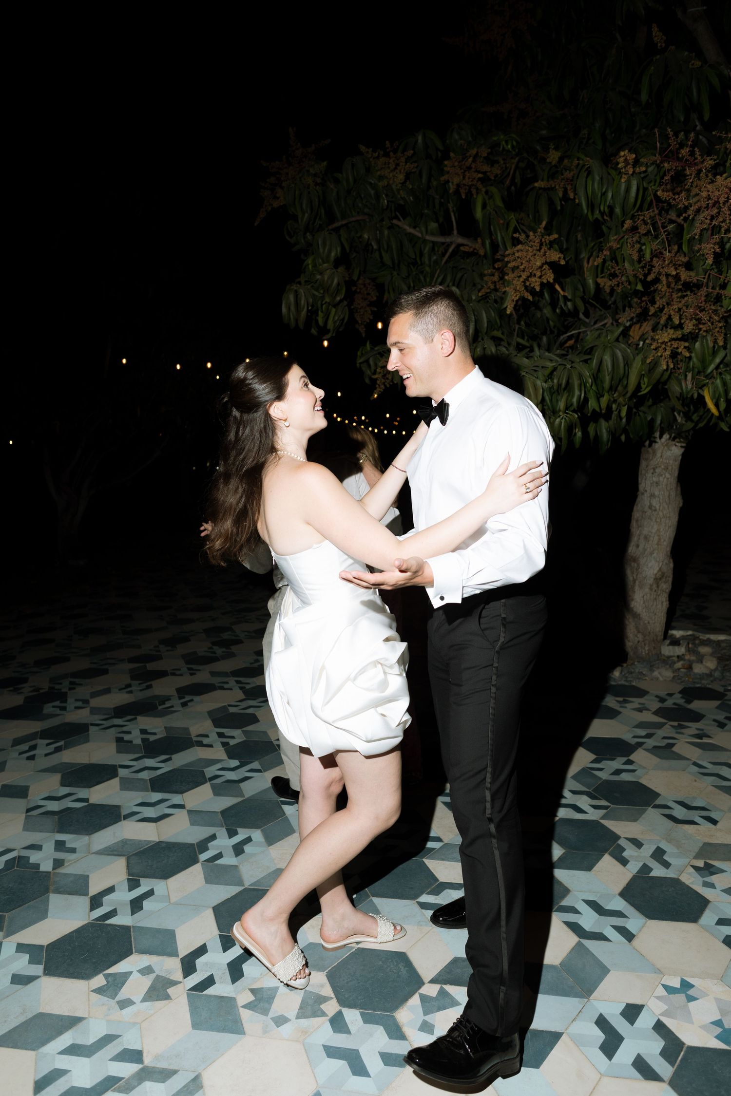 Bride in white mini dress and groom in tuxedo share a joyful first dance on a patterned outdoor floor at night.