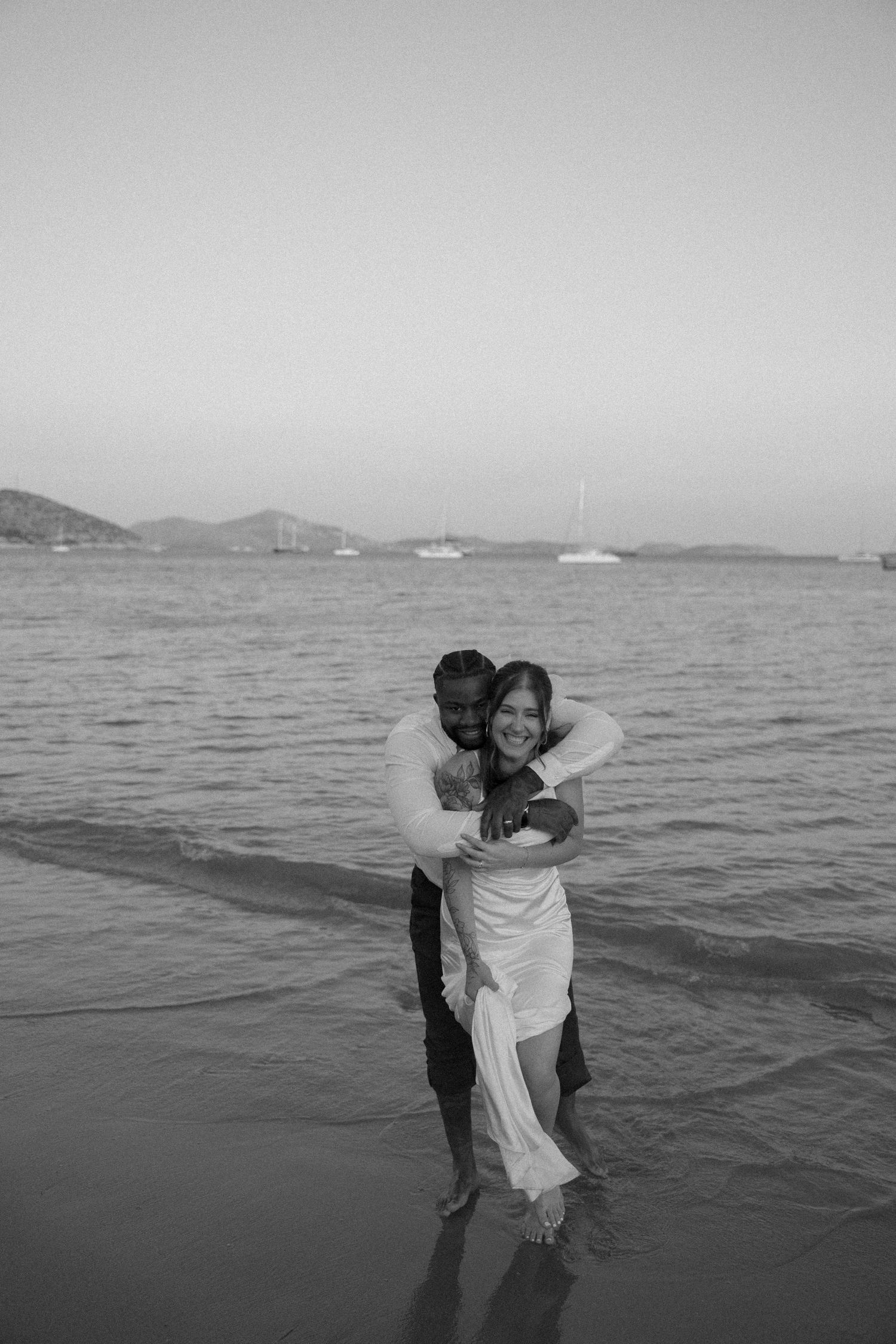 A couple embraces in black and white photography by the ocean with boats visible in the distance.