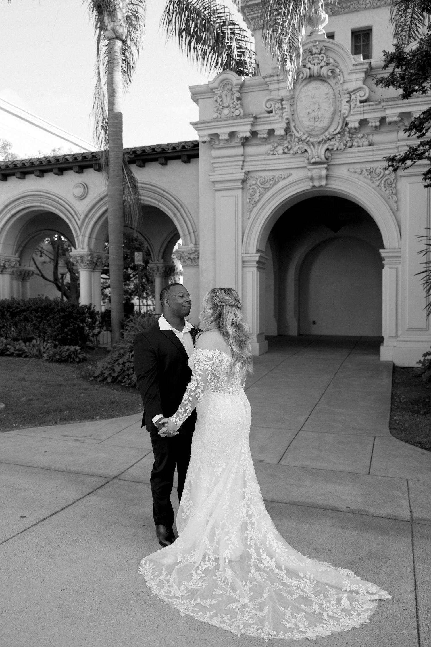 A couple in wedding attire poses in front of an ornate Spanish colonial style archway with palm trees.