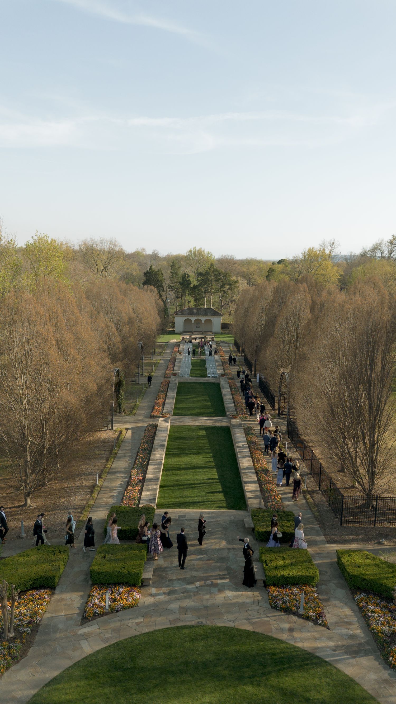 Aerial view of wedding guests walking through formal garden with manicured hedges and tree-lined pathway.