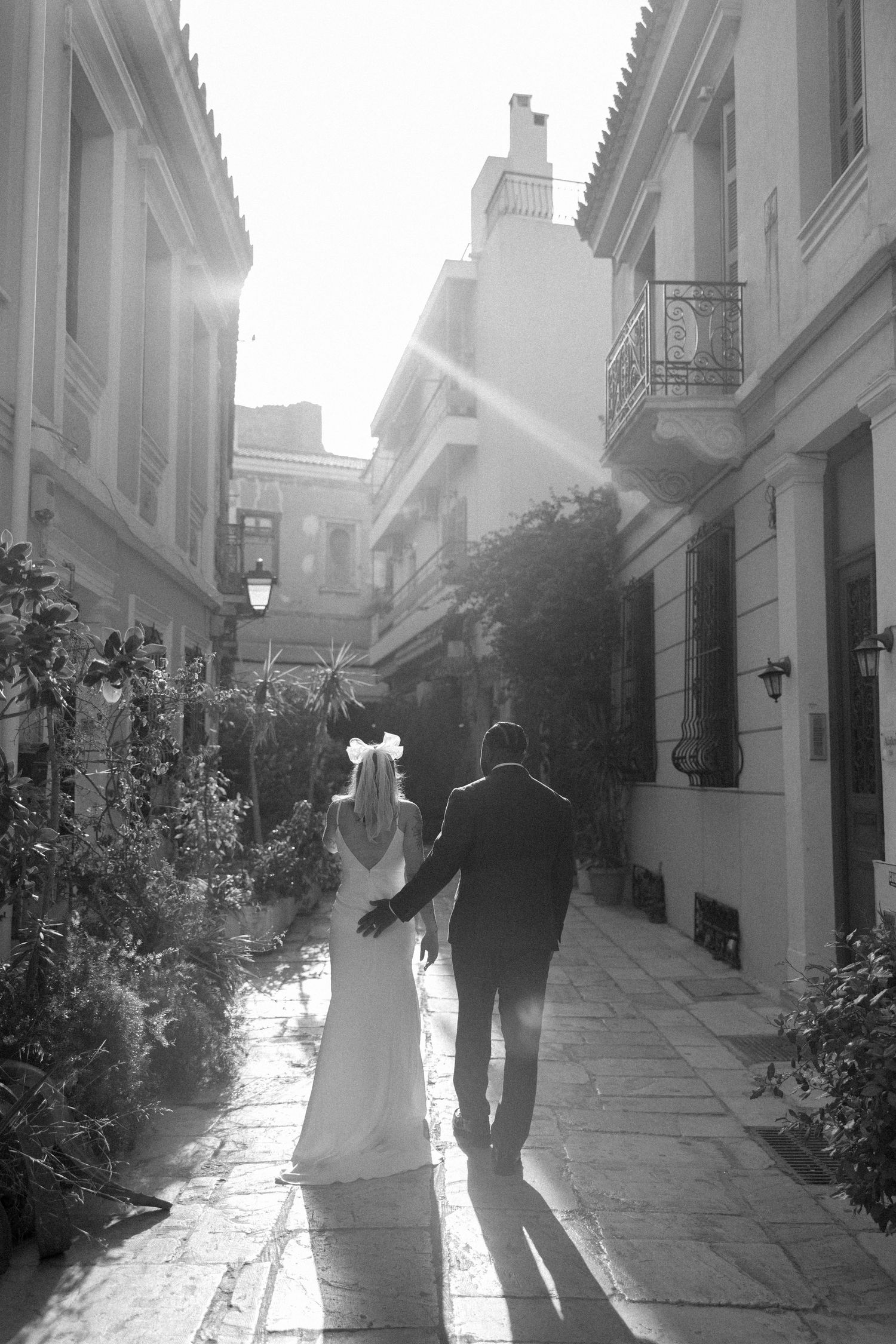 Black and white image of couple walking into sunlight on cobblestone path between buildings.