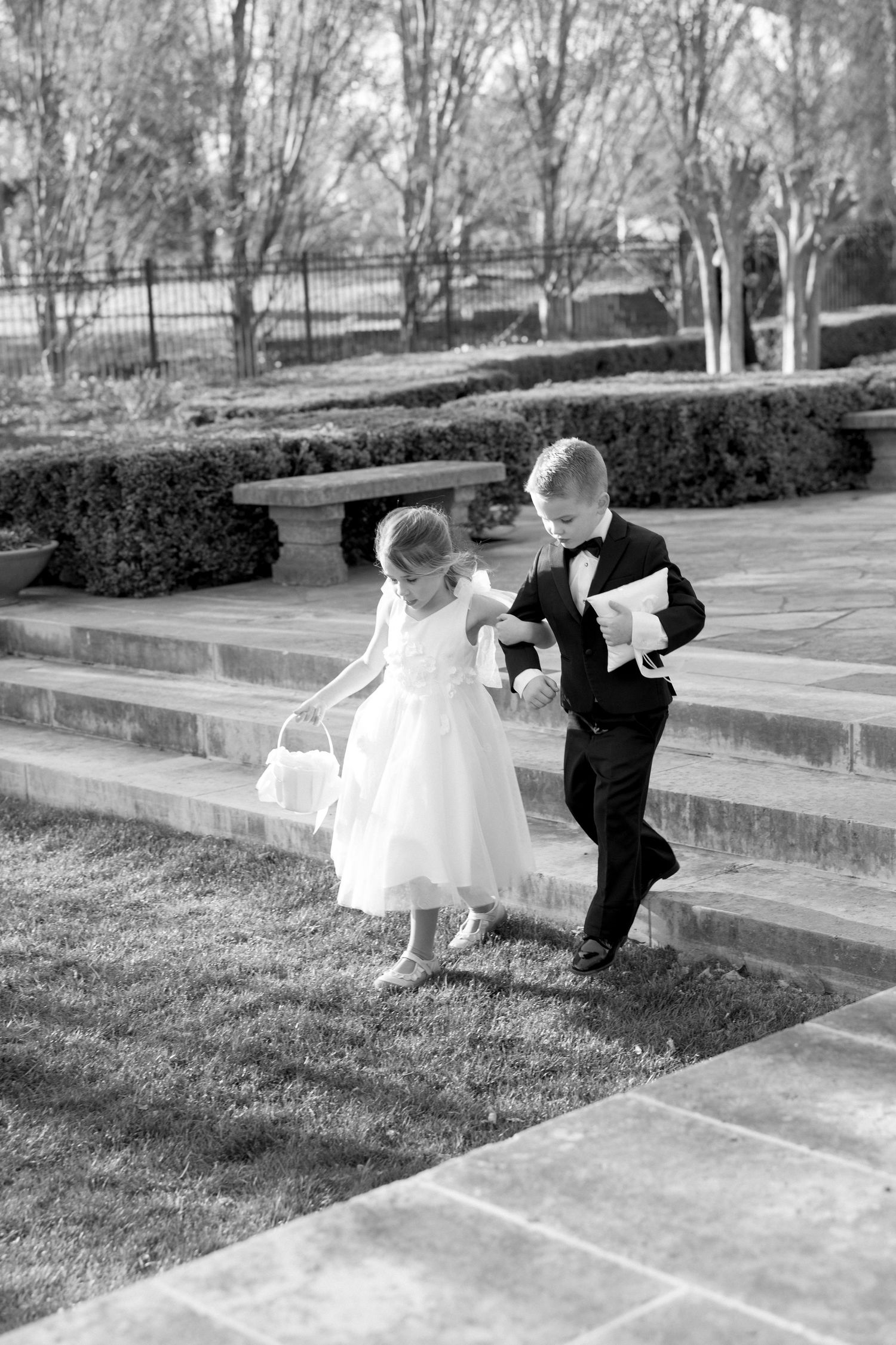 Flower girl and ring bearer in formal attire walk up stone steps at outdoor wedding, black and white.